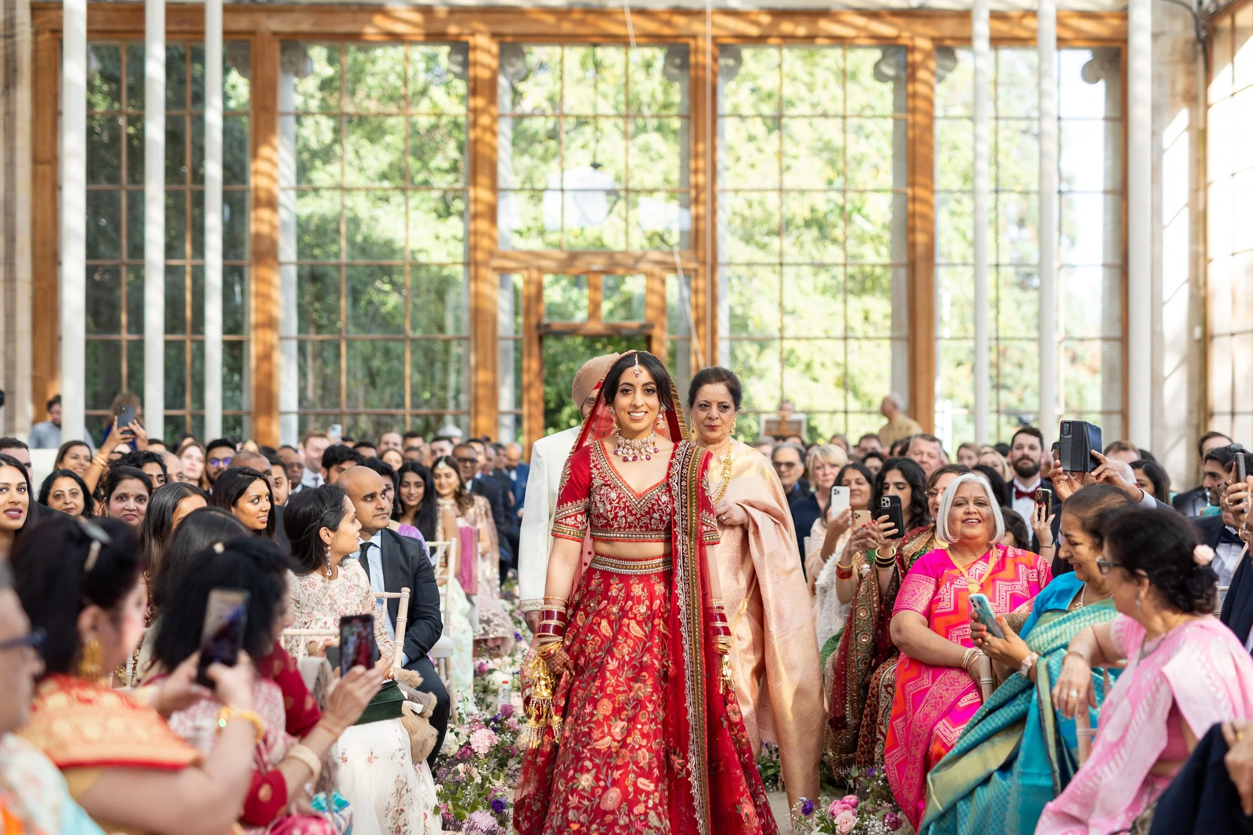 A bride in a red and gold traditional Indian bridal outfit walking down the aisle with a woman in a beige outfit, surrounded by seated and standing guests taking photos during a wedding ceremony in a sunny, glass-walled venue.