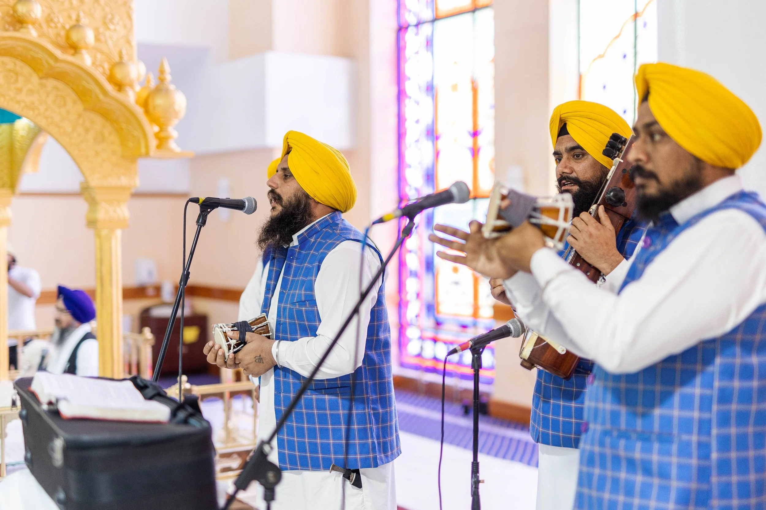 Three Sikh men wearing yellow turbans and blue plaid vests are performing Indian music with instruments and microphones in a decorated hall with stained glass windows.