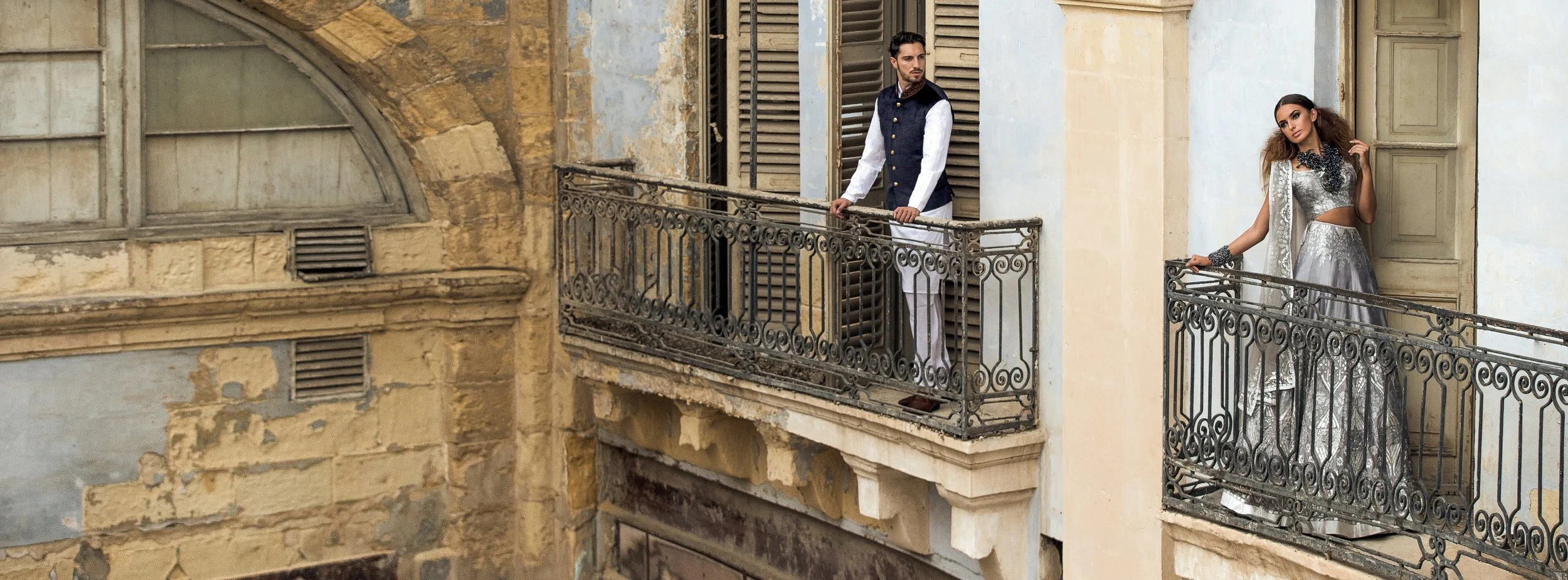 A man and woman standing on separate balconies of an old building with weathered walls. The man is gazing to the side, wearing a navy vest over a white shirt and white pants. The woman, dressed in a silver metallic gown with intricate lace details, is leaning on the balcony railing, looking at the camera. The building has a vintage appearance with chipped paint, wooden shutters, and decorative iron railings.