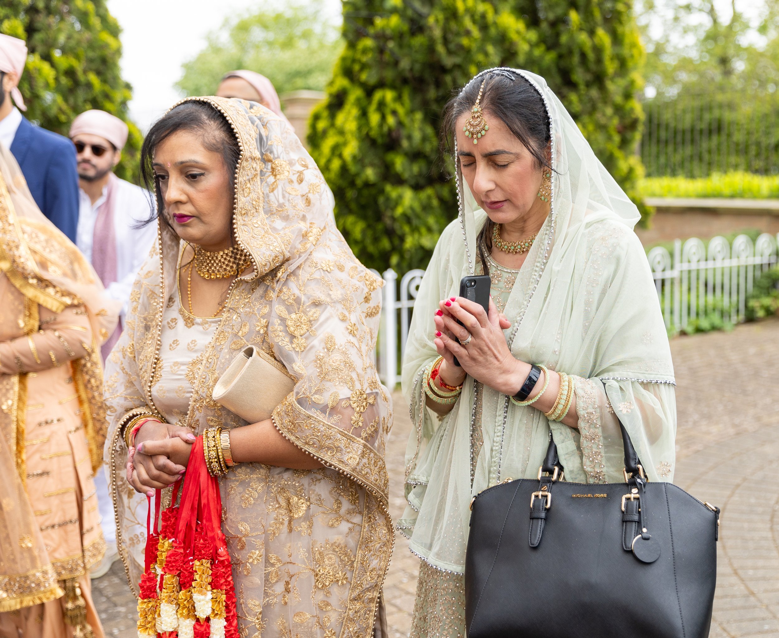 Two women in traditional Indian attire standing outdoors during a cultural event. One woman looks at her phone, while the other has her hands clasped and is holding a red and yellow garland.
