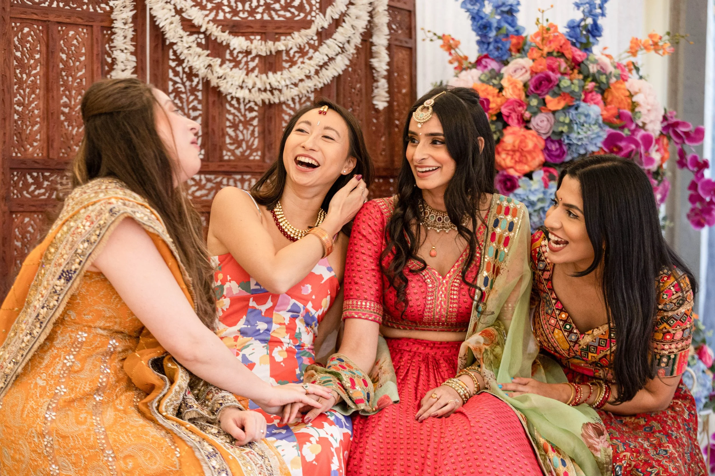 Four women of diverse ethnicities sitting together, dressed in colorful traditional Indian sarees and jewelry, enjoying a lively conversation at an indoor celebration with floral decorations and a wooden panel backdrop.