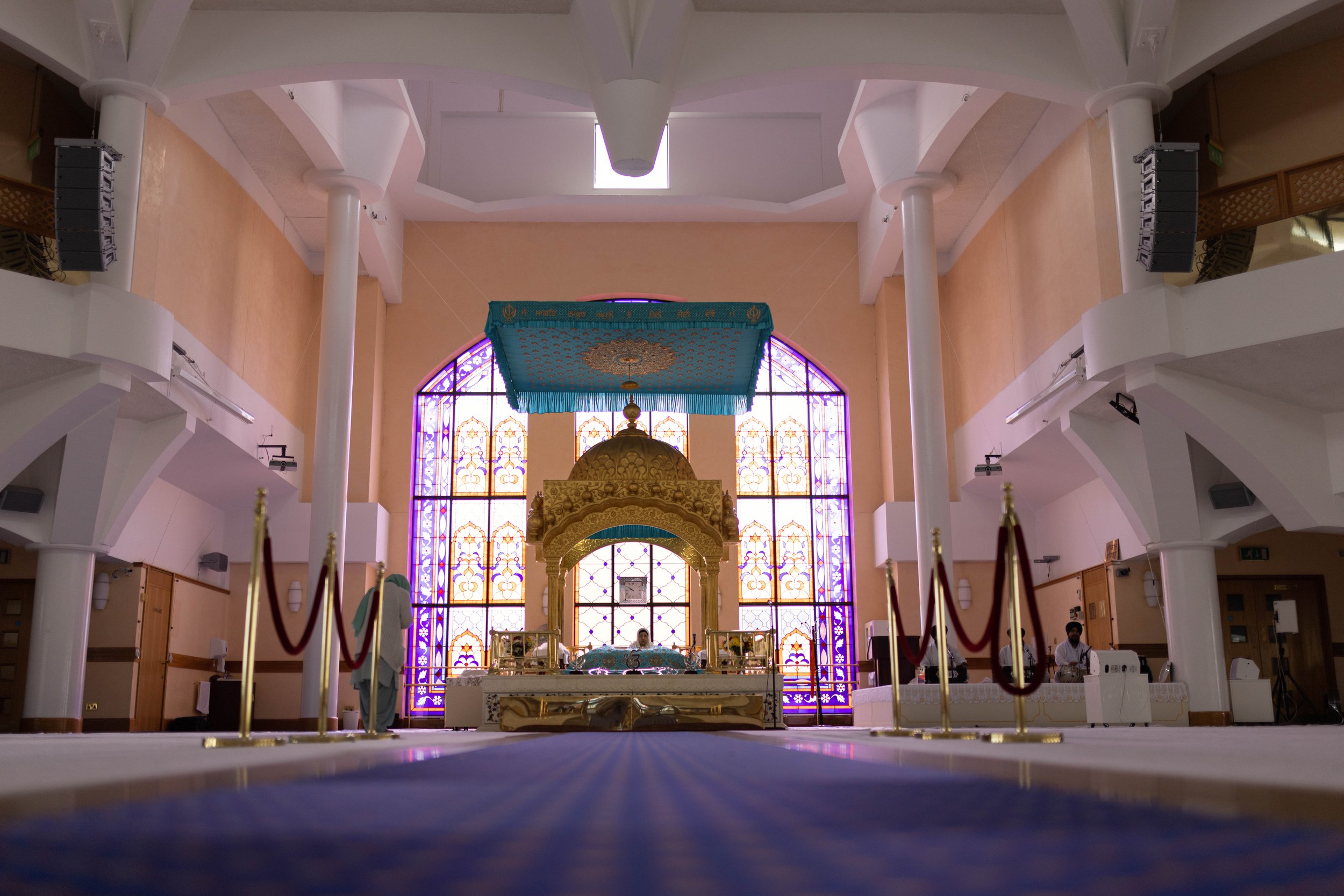 Interior of a Hindu temple with ornate golden shrine, large stained glass windows, and velvet canopy overhead.