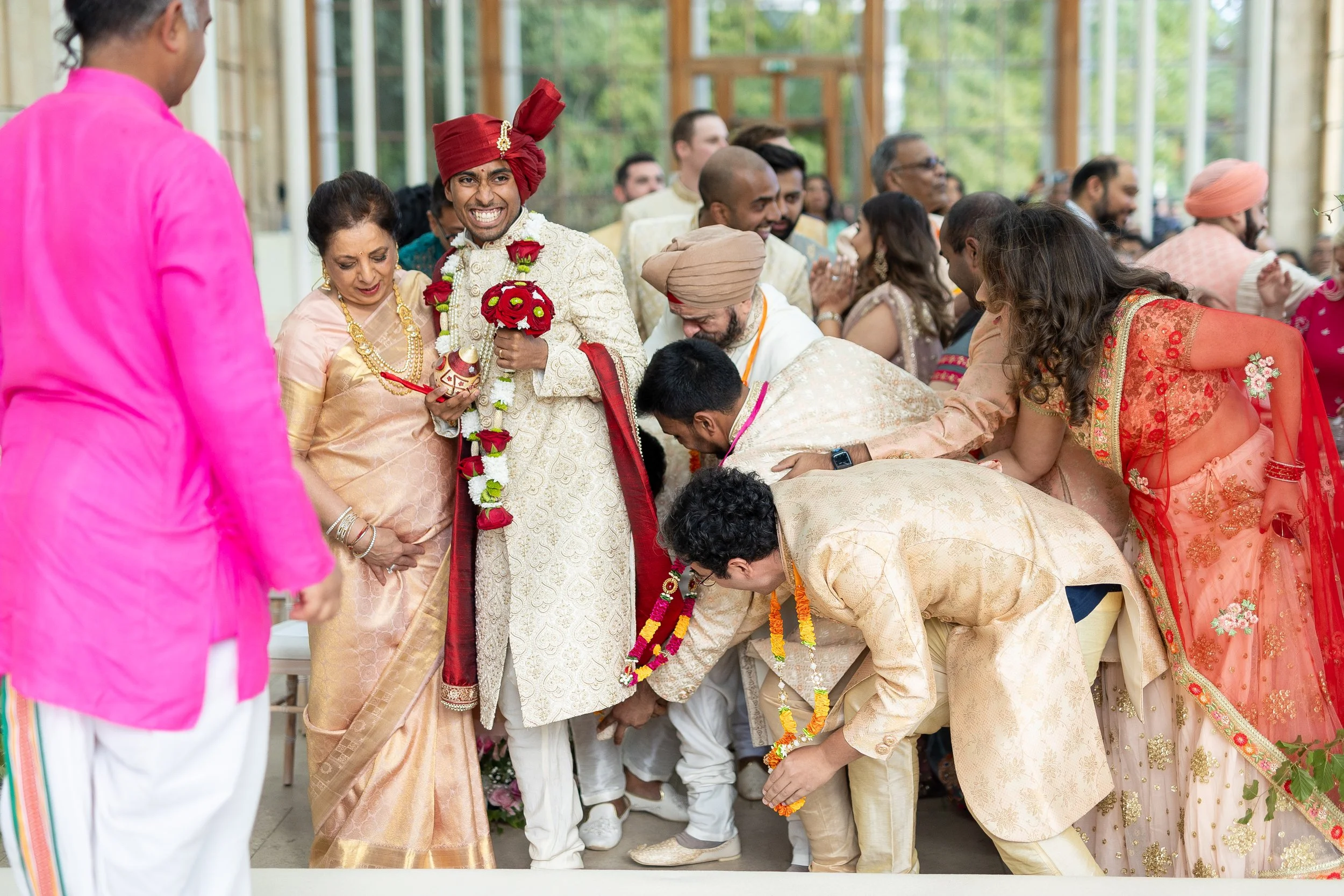 Indian wedding ceremony with the bride and groom dressed in traditional attire, surrounded by family and friends. The groom holds red roses, and guests bow down in a traditional gesture.