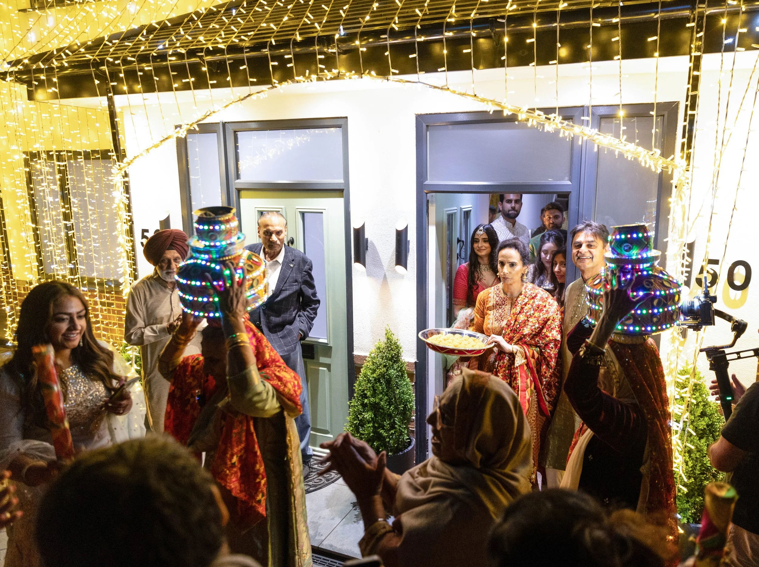 Indian family celebrating a festive occasion outside a house decorated with string lights, with two performers wearing colorful, illuminated costumes entertaining the guests.