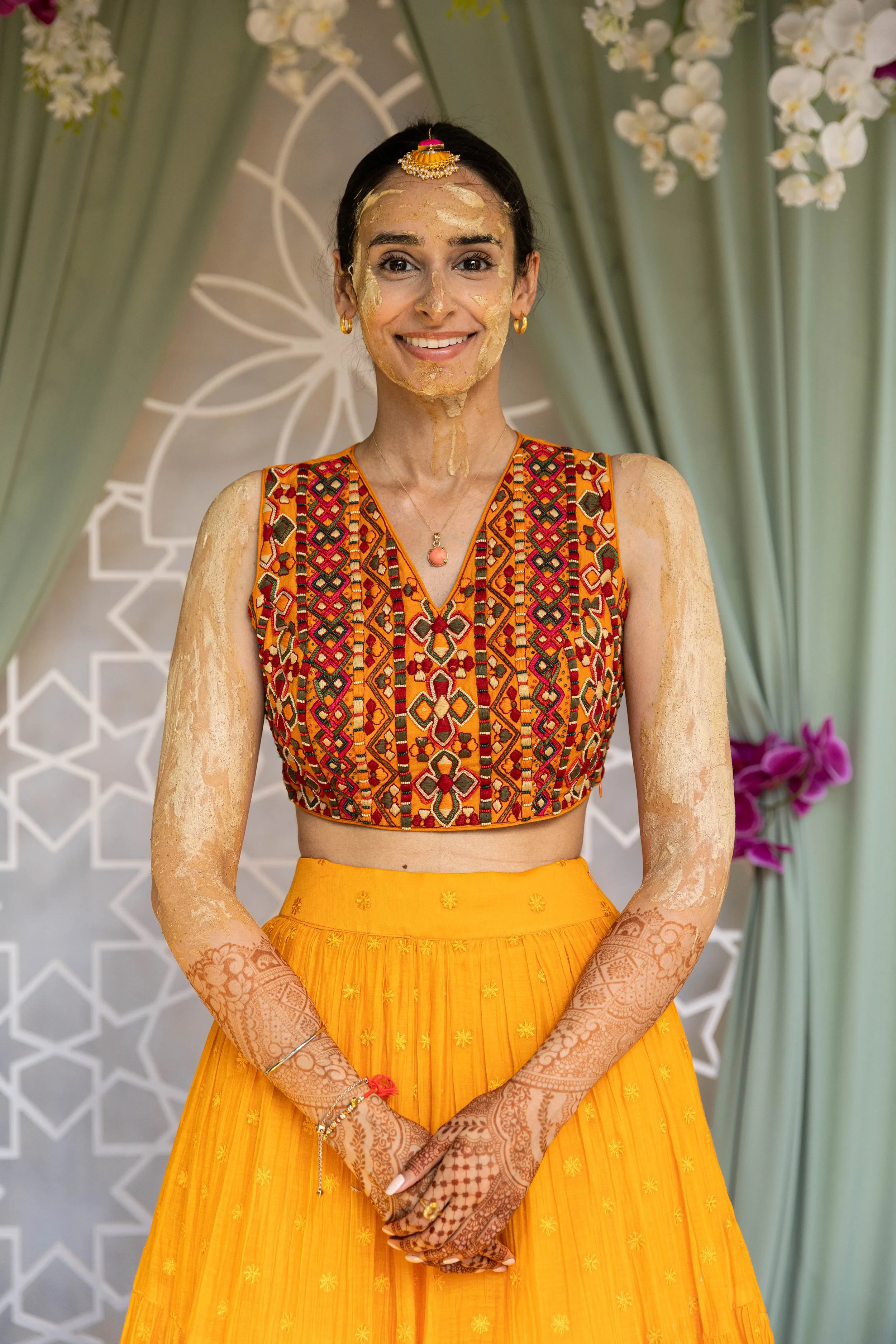 A woman in colorful traditional attire, smiling, with henna on her hands, standing in front of a decorative background with curtains, flowers, and geometric patterns.