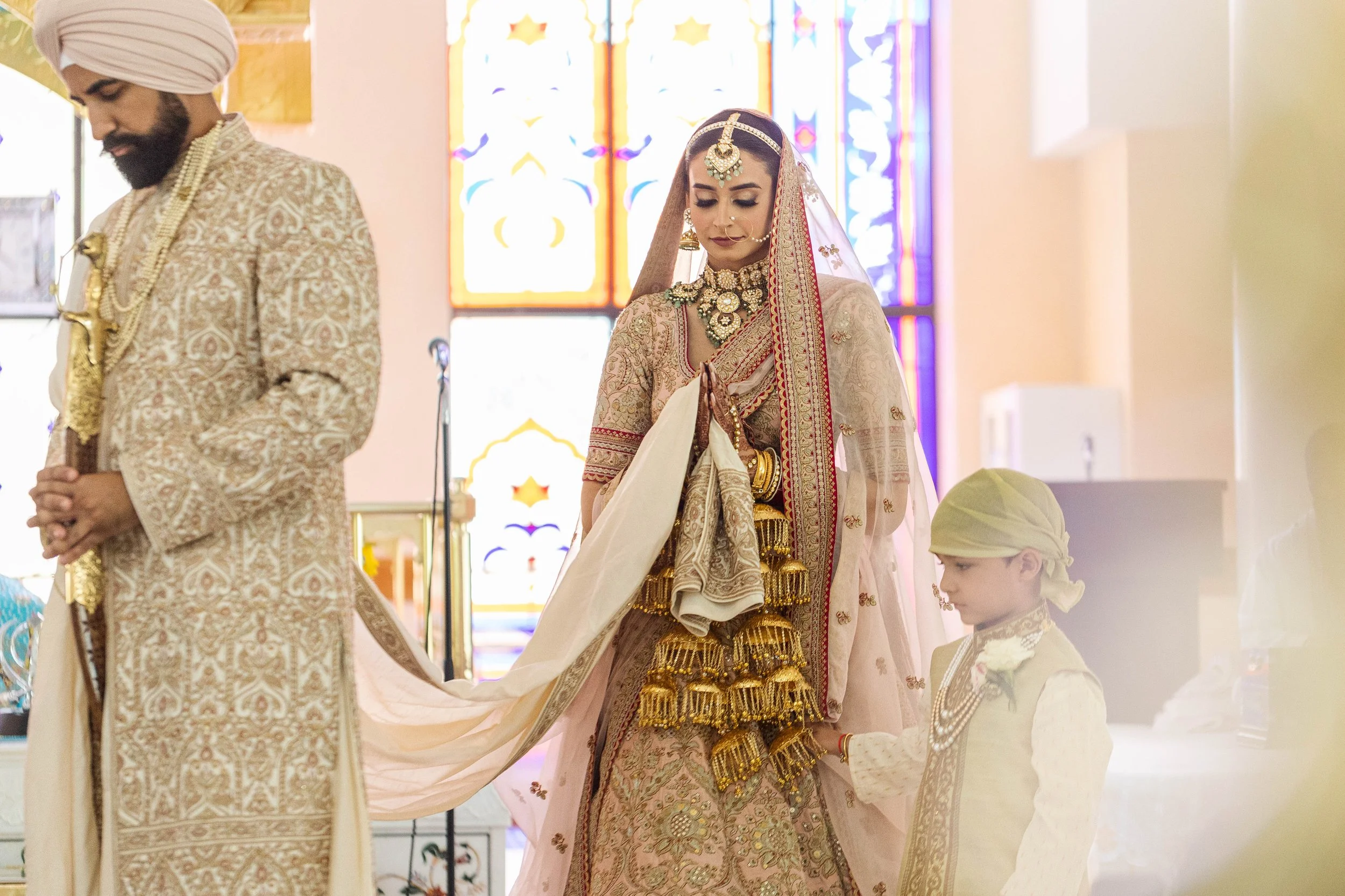 Indian bride dressed in traditional wedding attire with jewelry, standing with her hands clasped, accompanied by a young boy in traditional outfit, in a decorated setting with colorful stained glass windows.