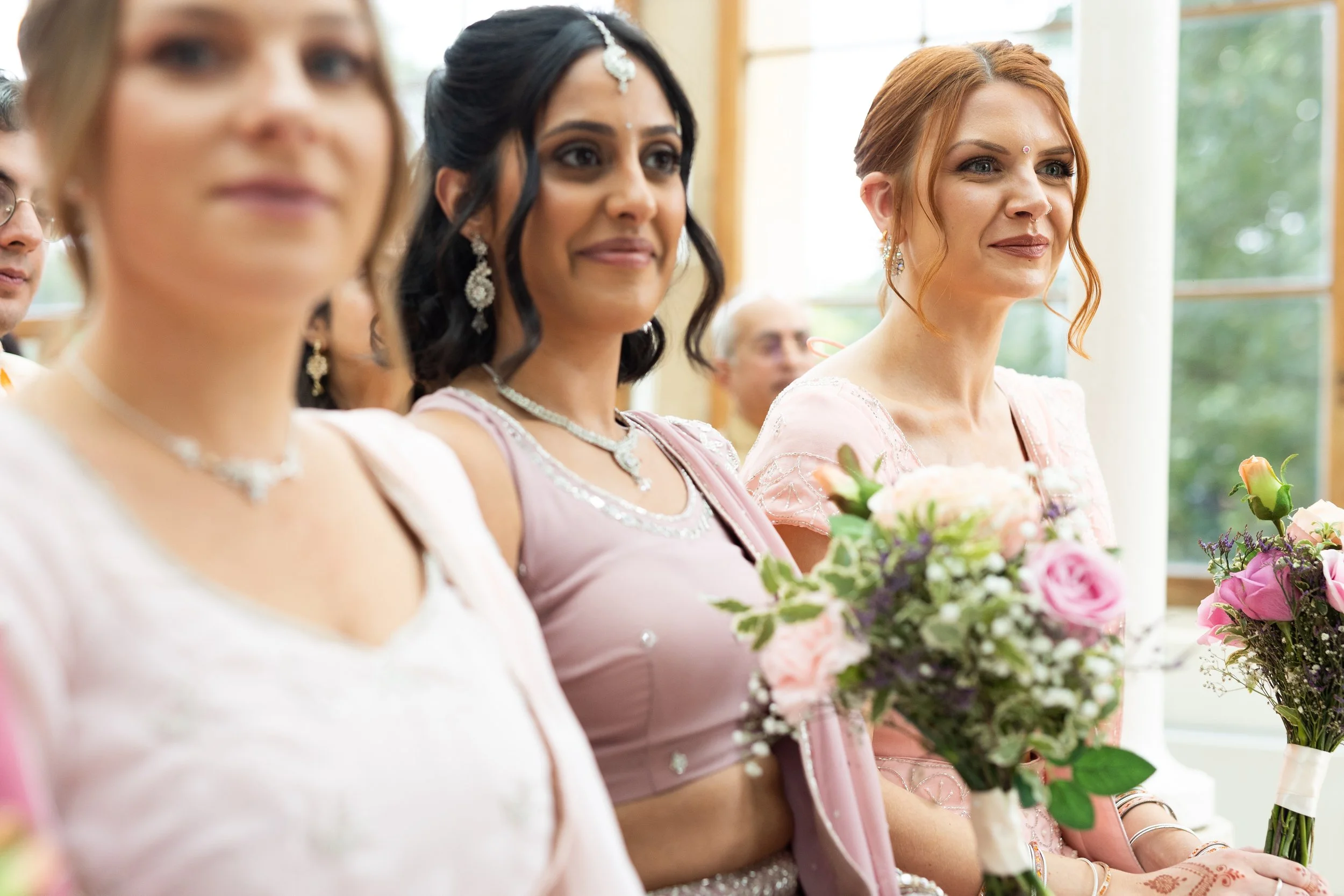 Three women sitting in a row holding floral bouquets, dressed in light-colored traditional attire, at a wedding ceremony.