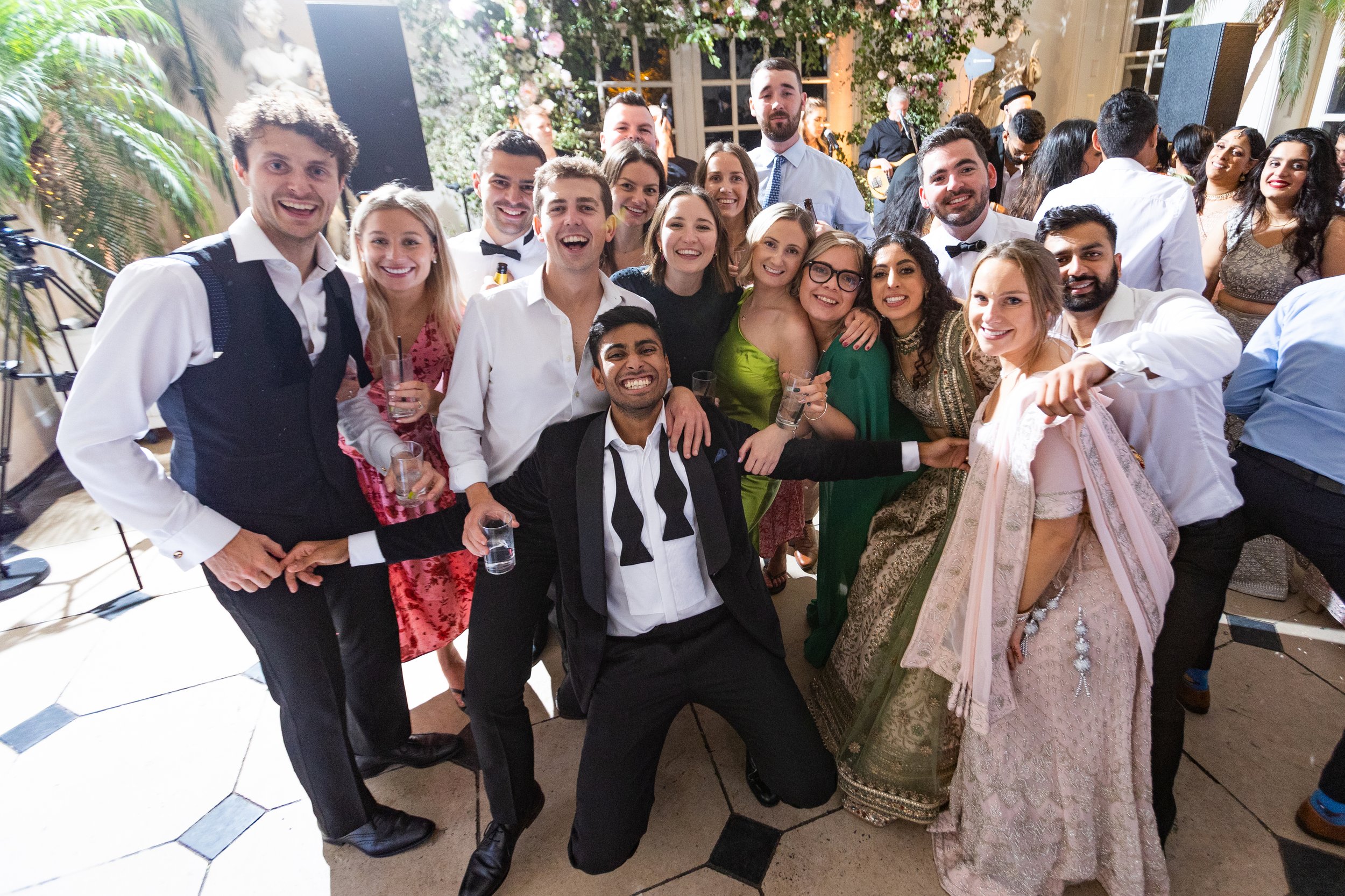A large group of people dressed in formal and traditional Indian attire, smiling and posing together at an indoor party or celebration.