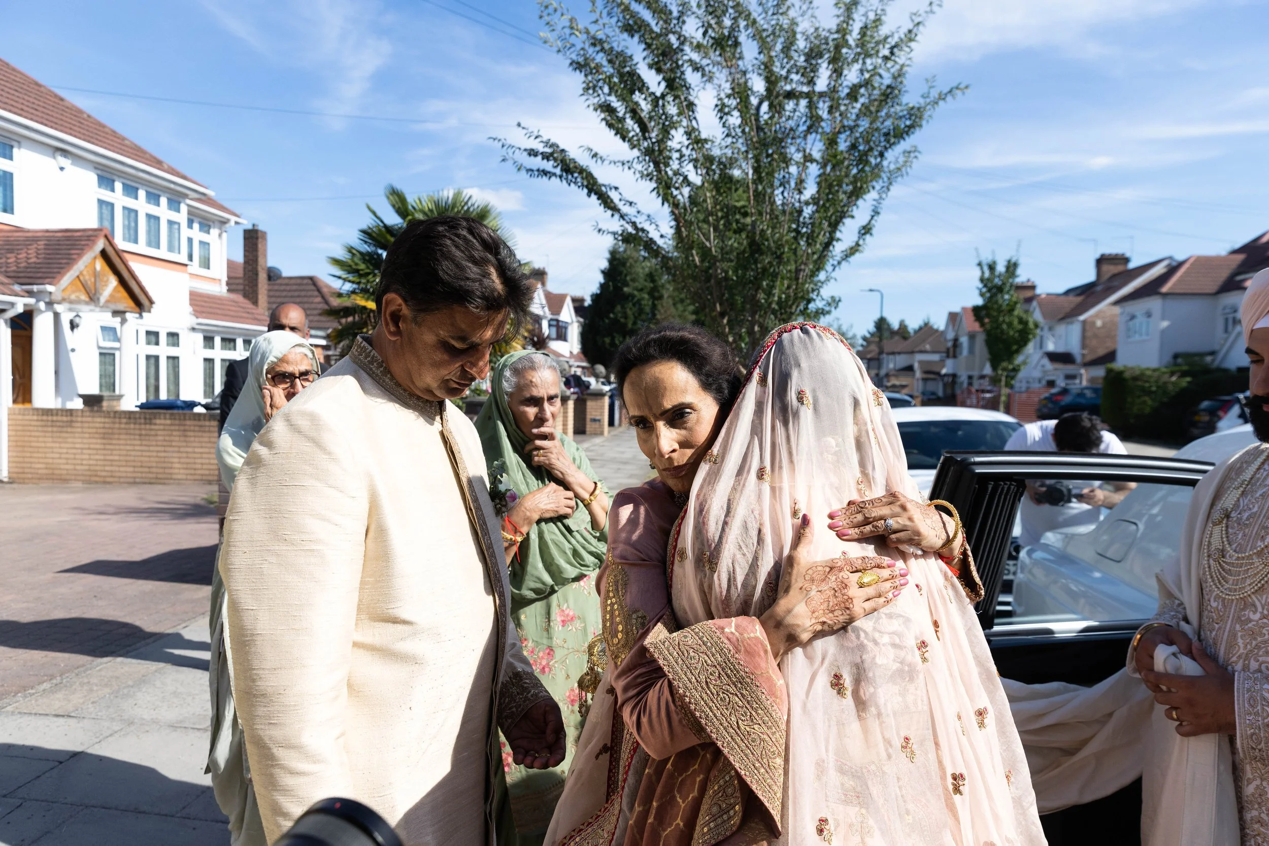 People dressed in traditional Indian clothing sharing a hug outdoors on a sunny residential street.