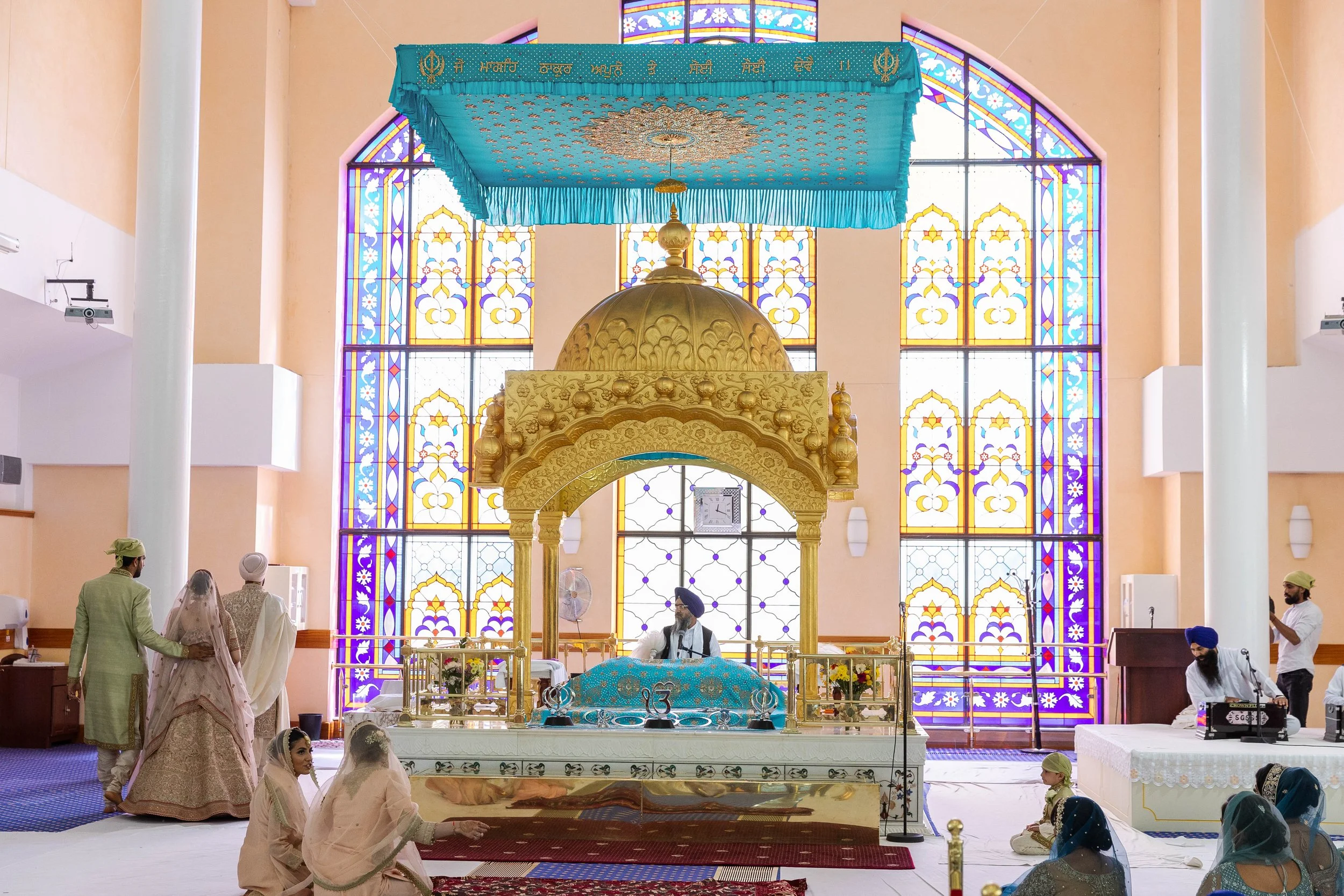 Interior of a Sikh Gurdwara with a gold throne and a man sitting on it, stained glass windows in the background, and several women and men seated on the floor.