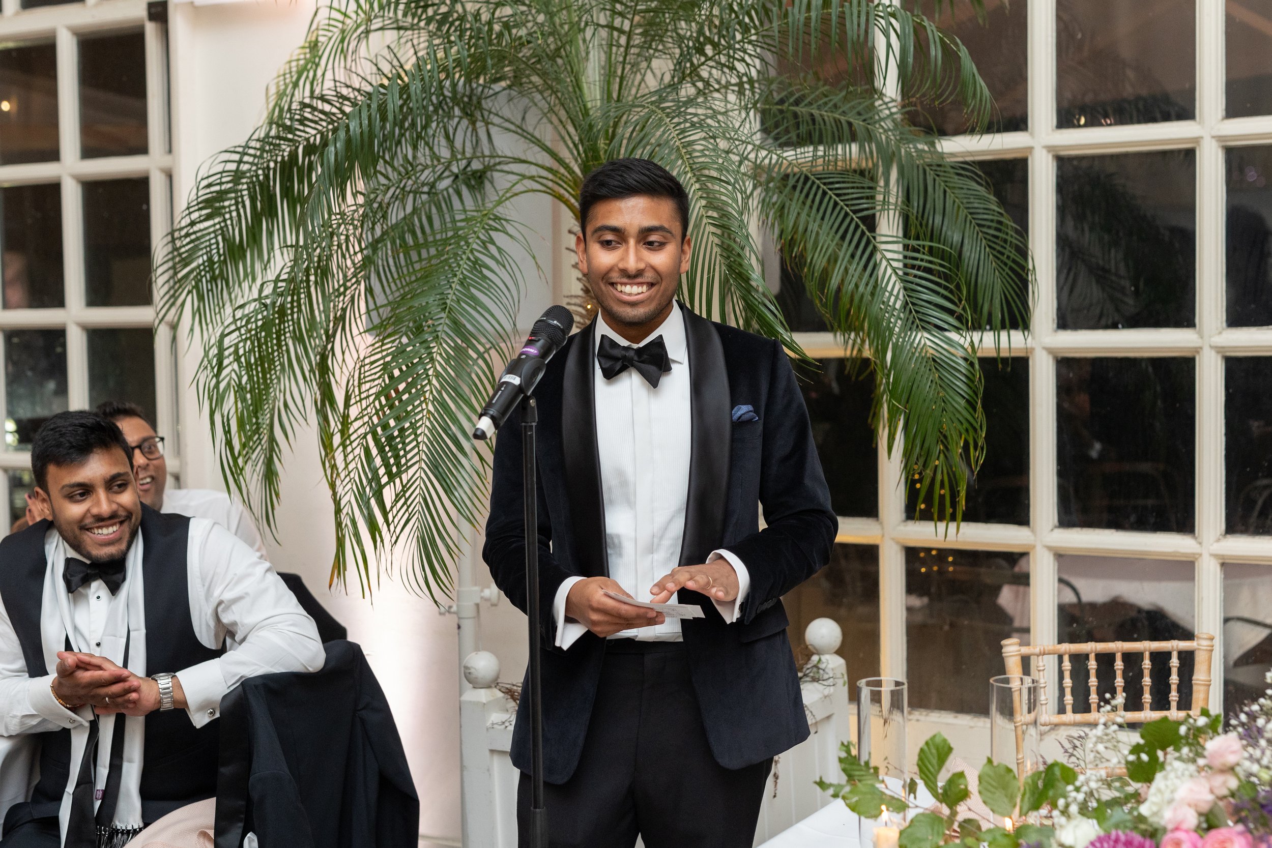 A man in a tuxedo giving a speech at a wedding reception, with a large green plant behind him and people sitting at a table, smiling and listening.