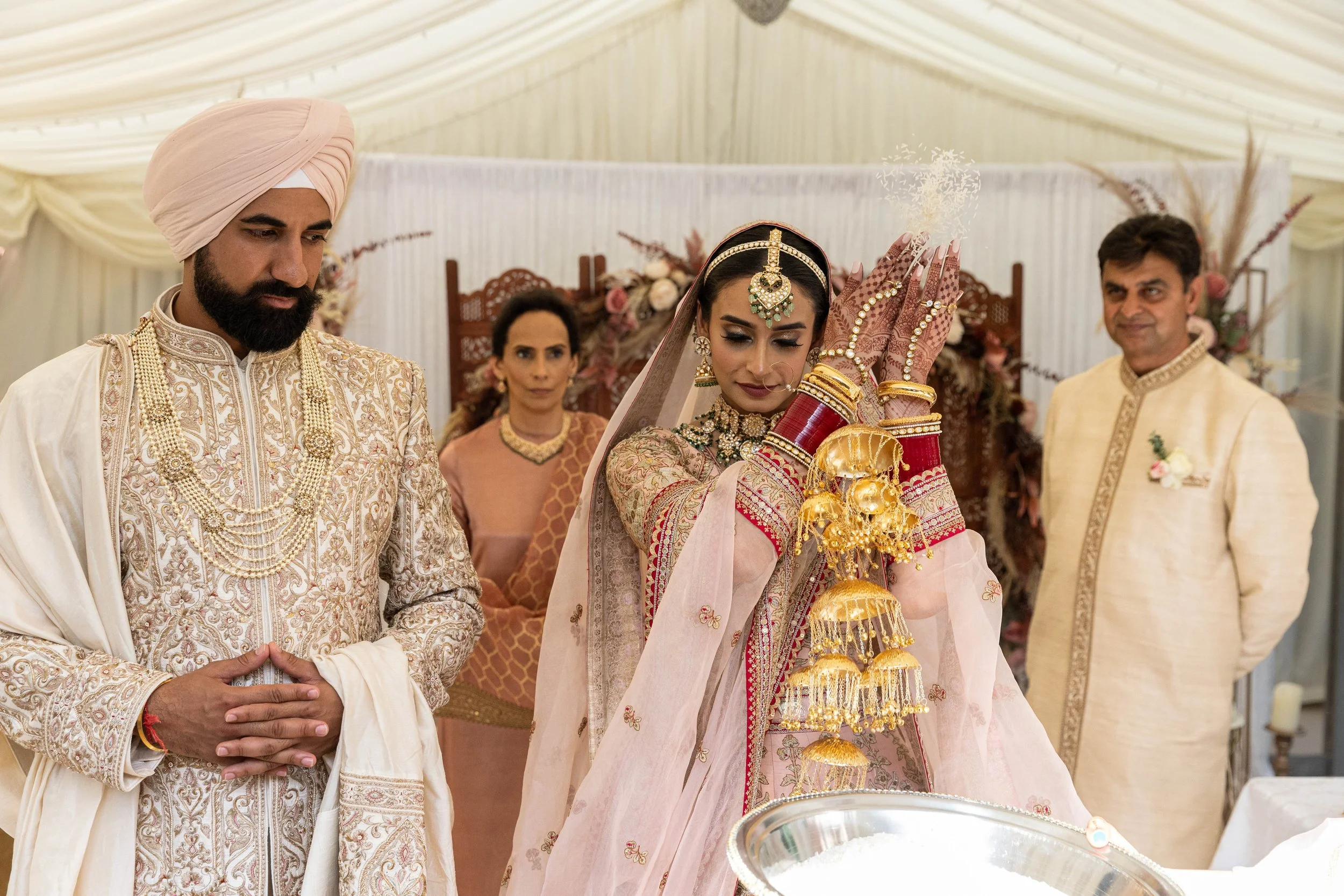 Indian bride performing a traditional ceremony with family members. The bride is dressed in a pink and gold lehenga with red and gold bangles, and jewelry, holding a ceremonial item. Family members, dressed in traditional attire, stand around her ins