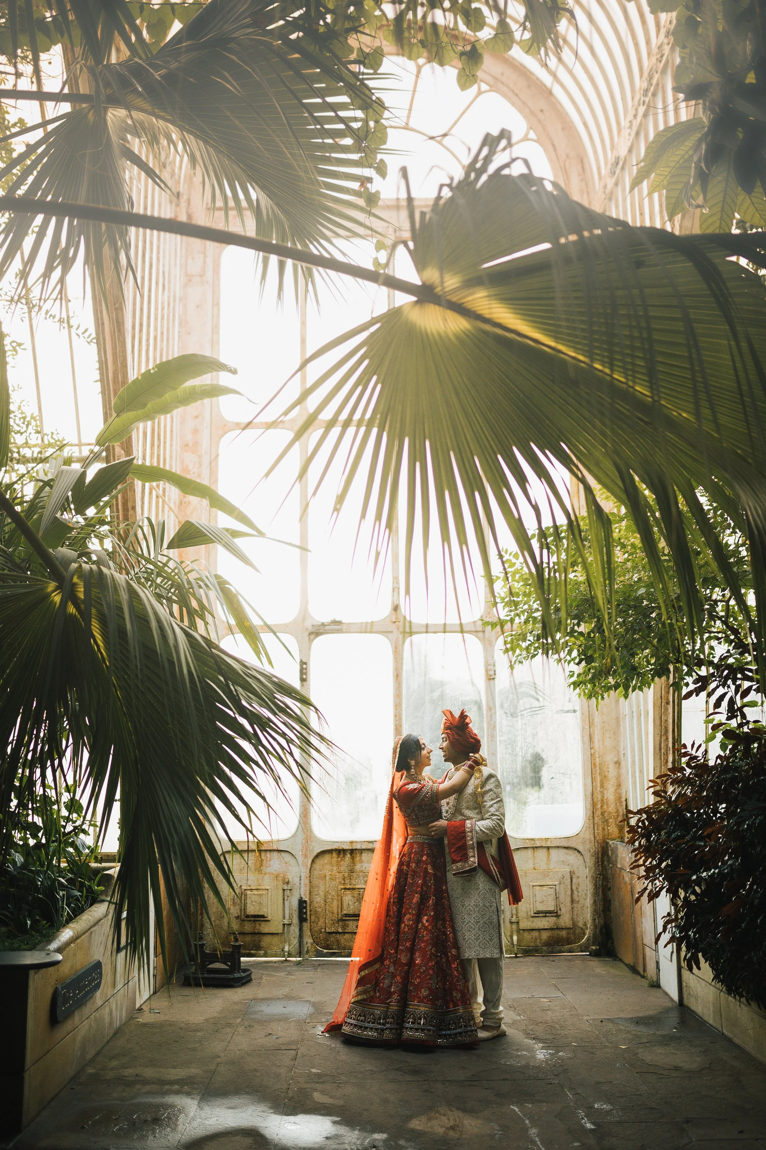 A couple dressed in traditional Indian wedding attire stands close together inside a lush greenhouse or conservatory with tropical plants and large glass windows.