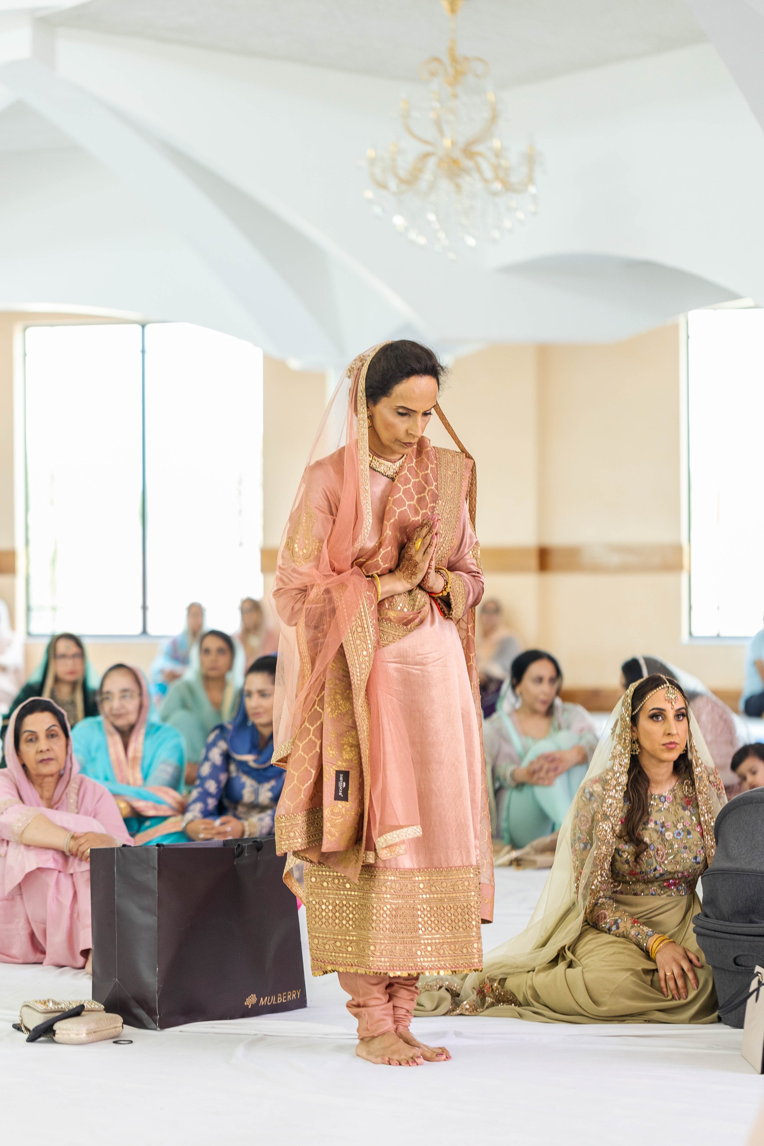 A woman in traditional South Asian attire praying with hands pressed together during a ceremony, surrounded by seated guests.