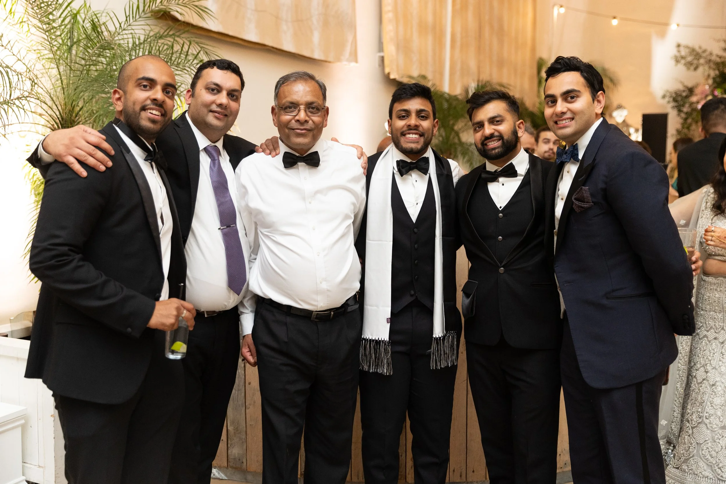 Group of seven men in formal attire, smiling at a celebration or wedding reception, with plants and warm lighting in the background.