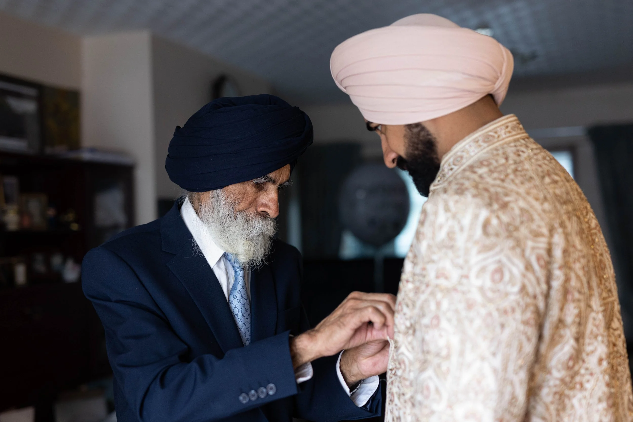 An elderly Sikh man dressing a younger Sikh man in traditional attire.