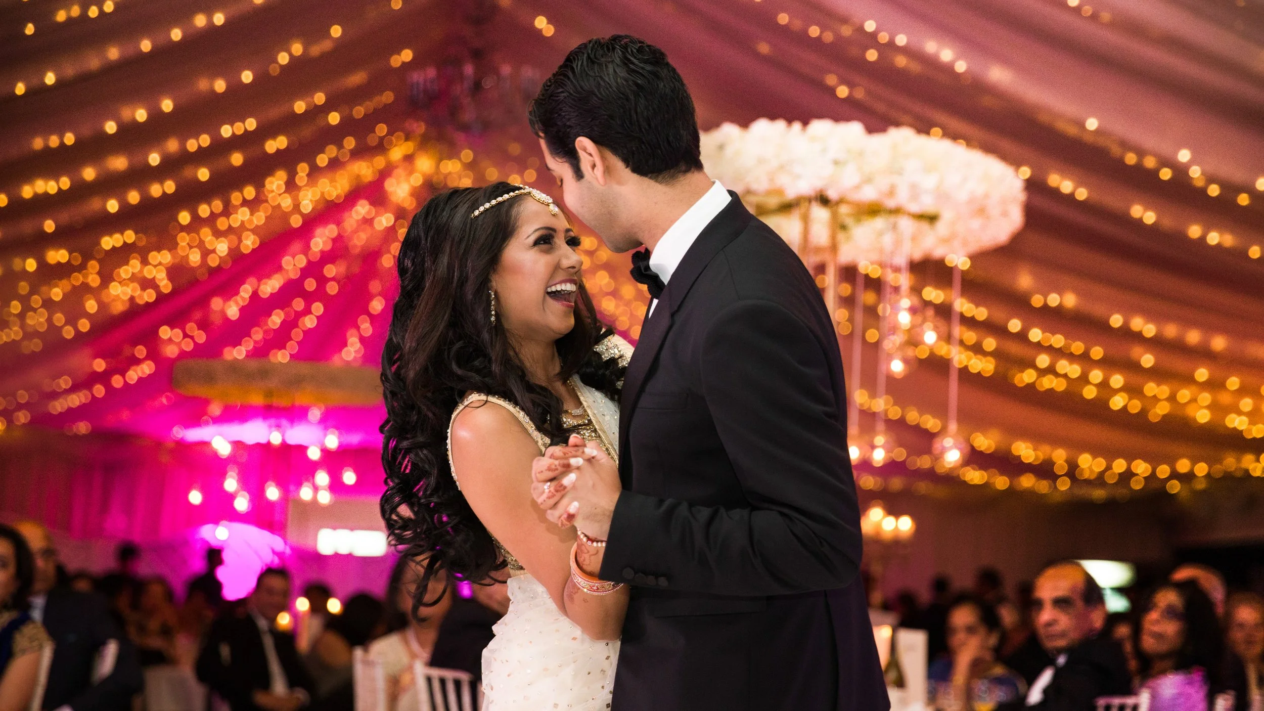 A bride and groom dance closely at their wedding reception, smiling and holding hands, with a decorated ceiling and warm lighting in the background.