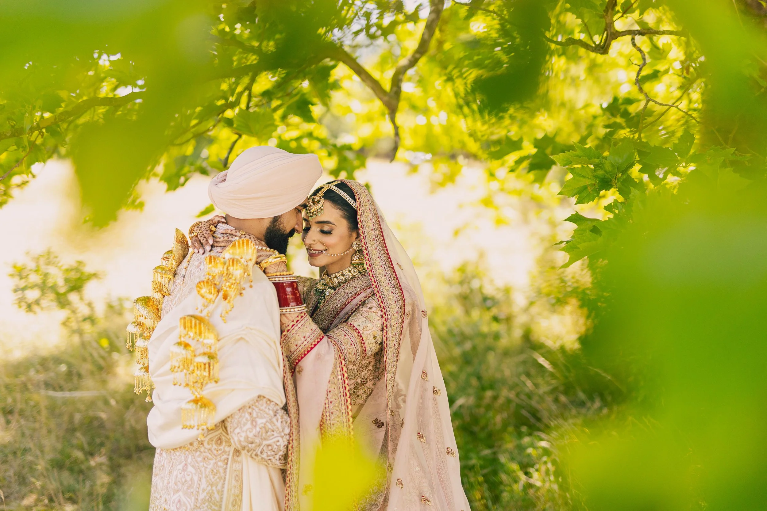 A bride and groom in traditional Indian wedding attire sharing a close moment outdoors, surrounded by green leaves and trees.