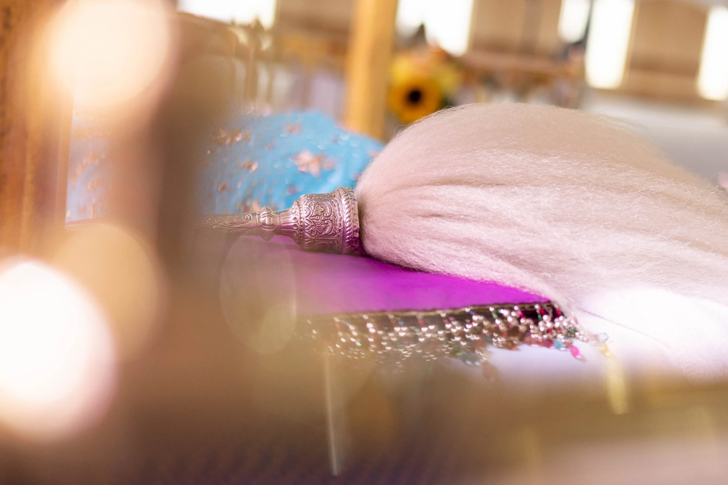Close-up of a pink feather duster with a silver handle resting on a purple surface. The background is blurred with warm lighting and suggests a decorative interior setting.