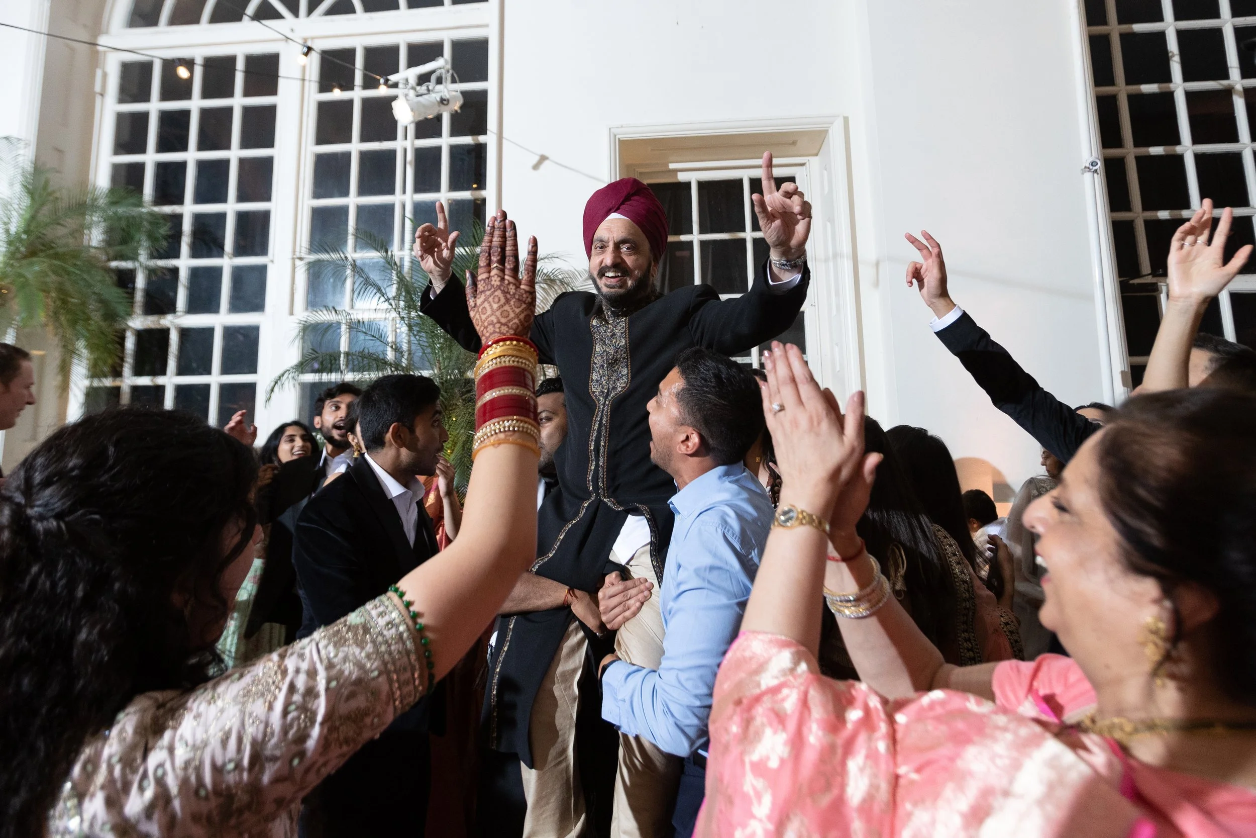 People dancing and celebrating at a festive event, with a man wearing a traditional turban being lifted in the air.