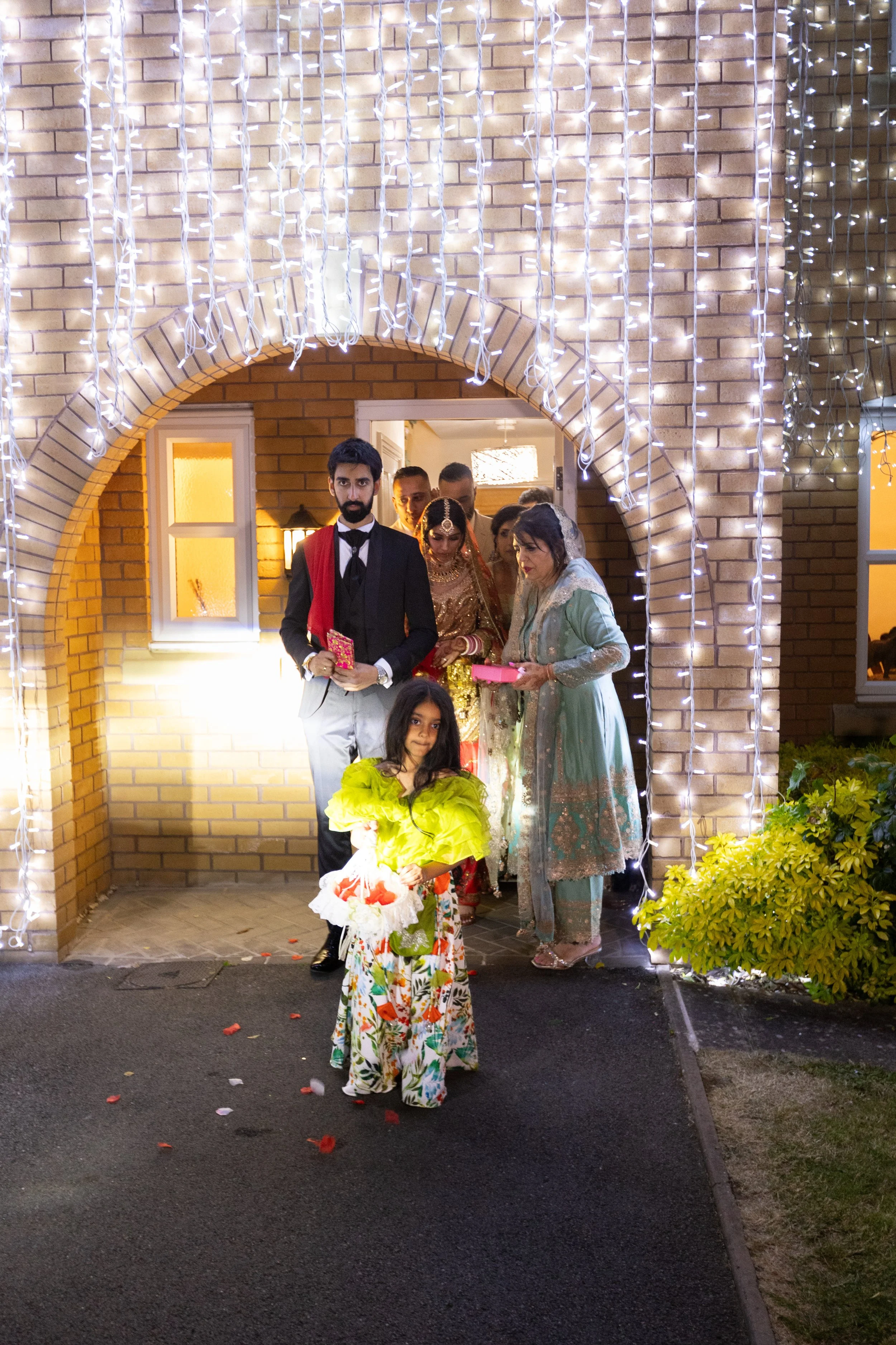 A group of people, including a young girl holding flowers, is standing outside a house decorated with white string lights, possibly celebrating a special occasion.