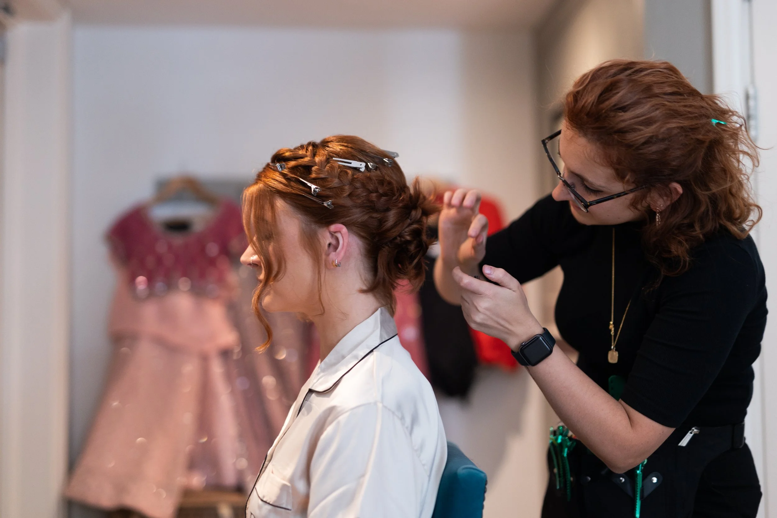 A stylist is helping a woman with her hair, which is styled into an updo with clips, in a dressing room with pink dresses hanging in the background.