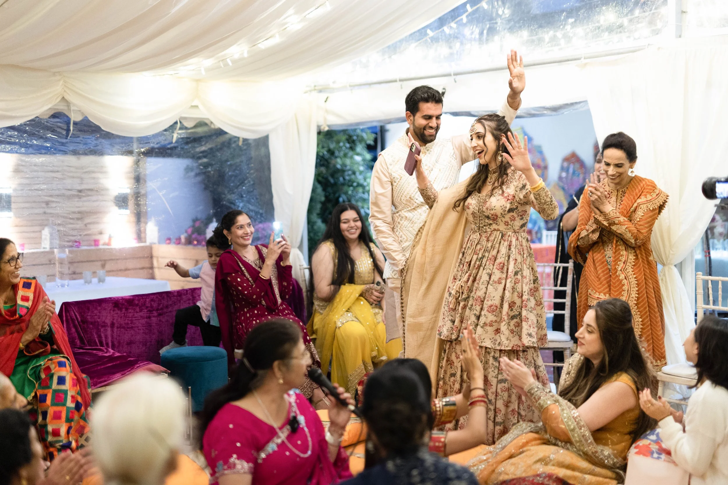 Group of people at a celebration, wearing traditional South Asian clothing, dancing and smiling under a decorated tent.