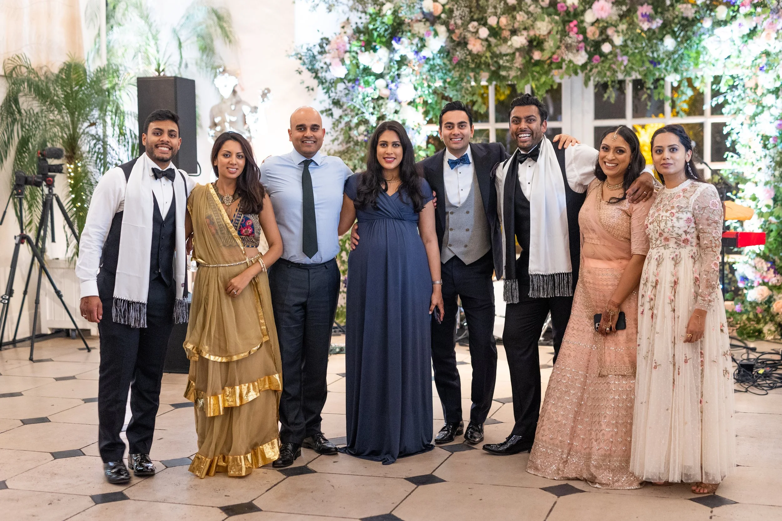 Group of nine people dressed in formal attire, standing together in front of a decorated floral background at a celebration or wedding event.
