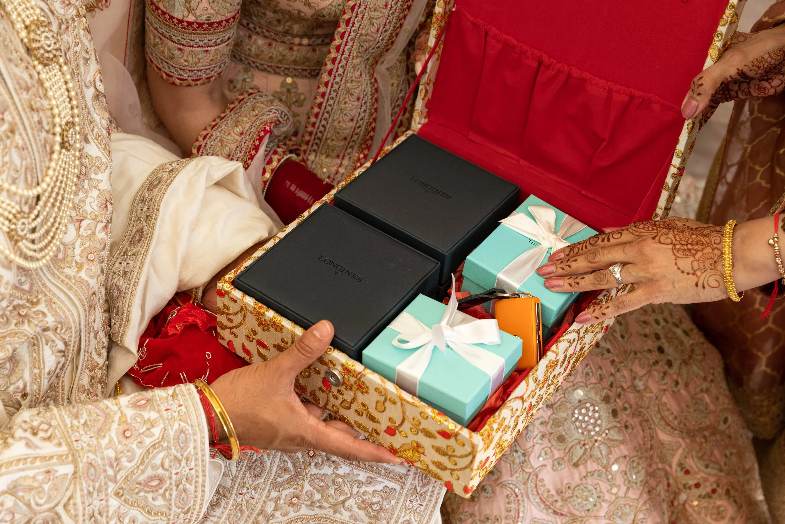 A person's hand holding an ornate jewelry box filled with gift-wrapped presents, including black Longines watches and small pastel gift boxes with white ribbons, during a celebration dressed in traditional Indian attire.