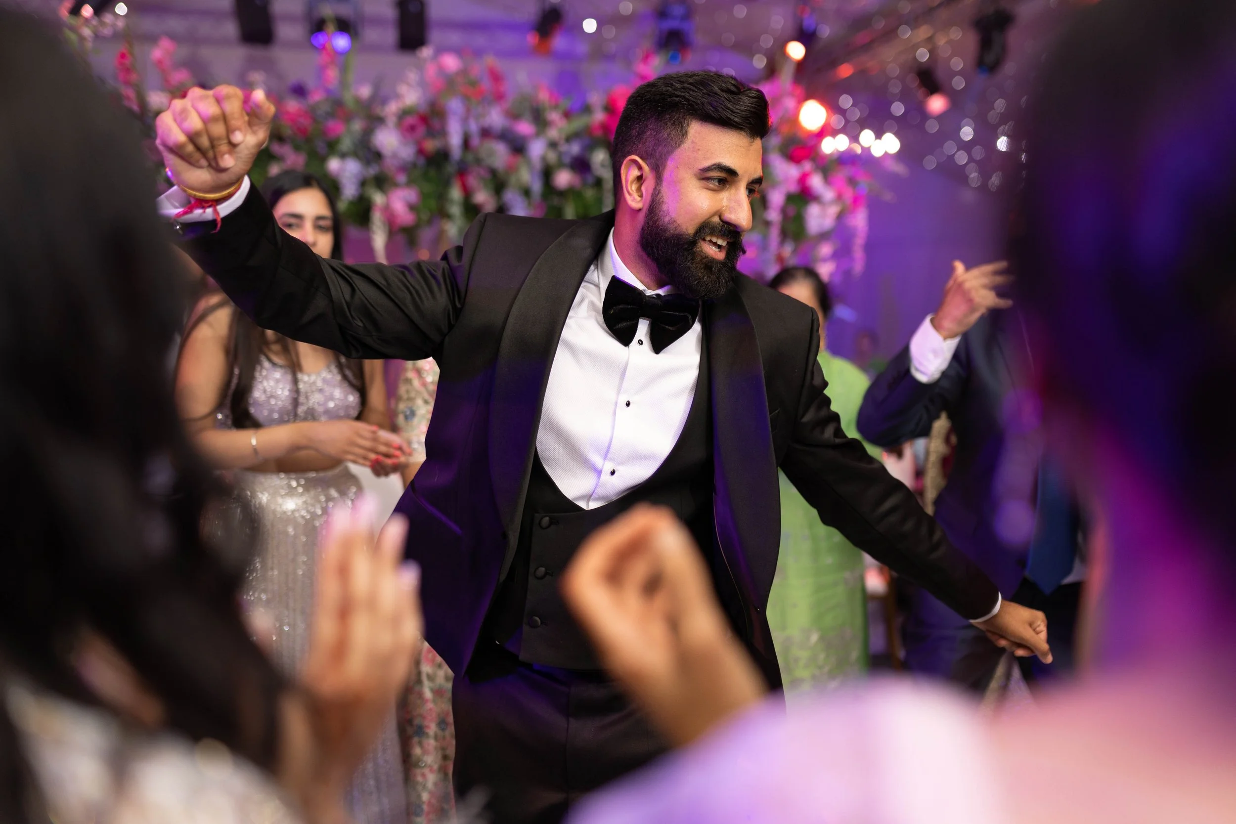 A man in a tuxedo and bow tie dancing and smiling at a celebration or wedding reception, with people and floral decorations in the background.
