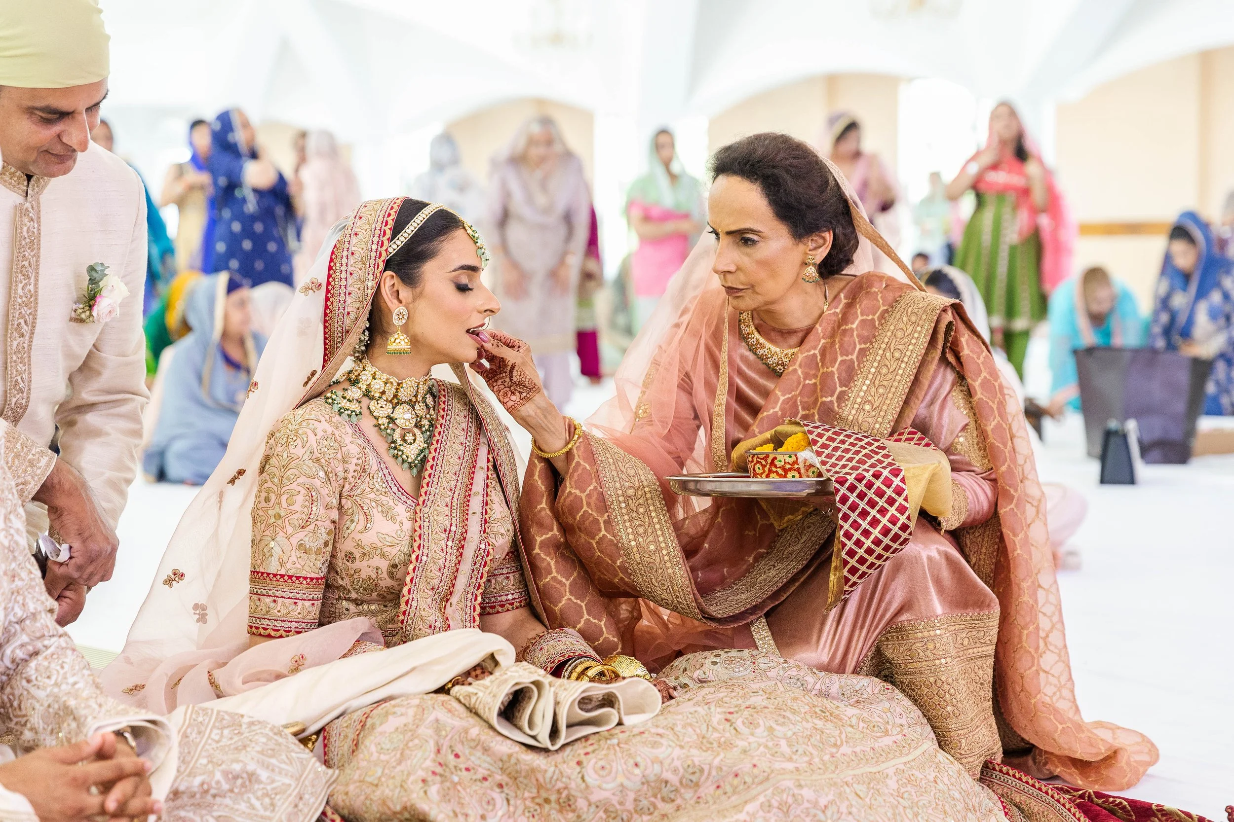 Indian bride in traditional attire participating in a wedding ritual, with an elder woman applying something to her lips, surrounded by family members in colorful traditional clothing.