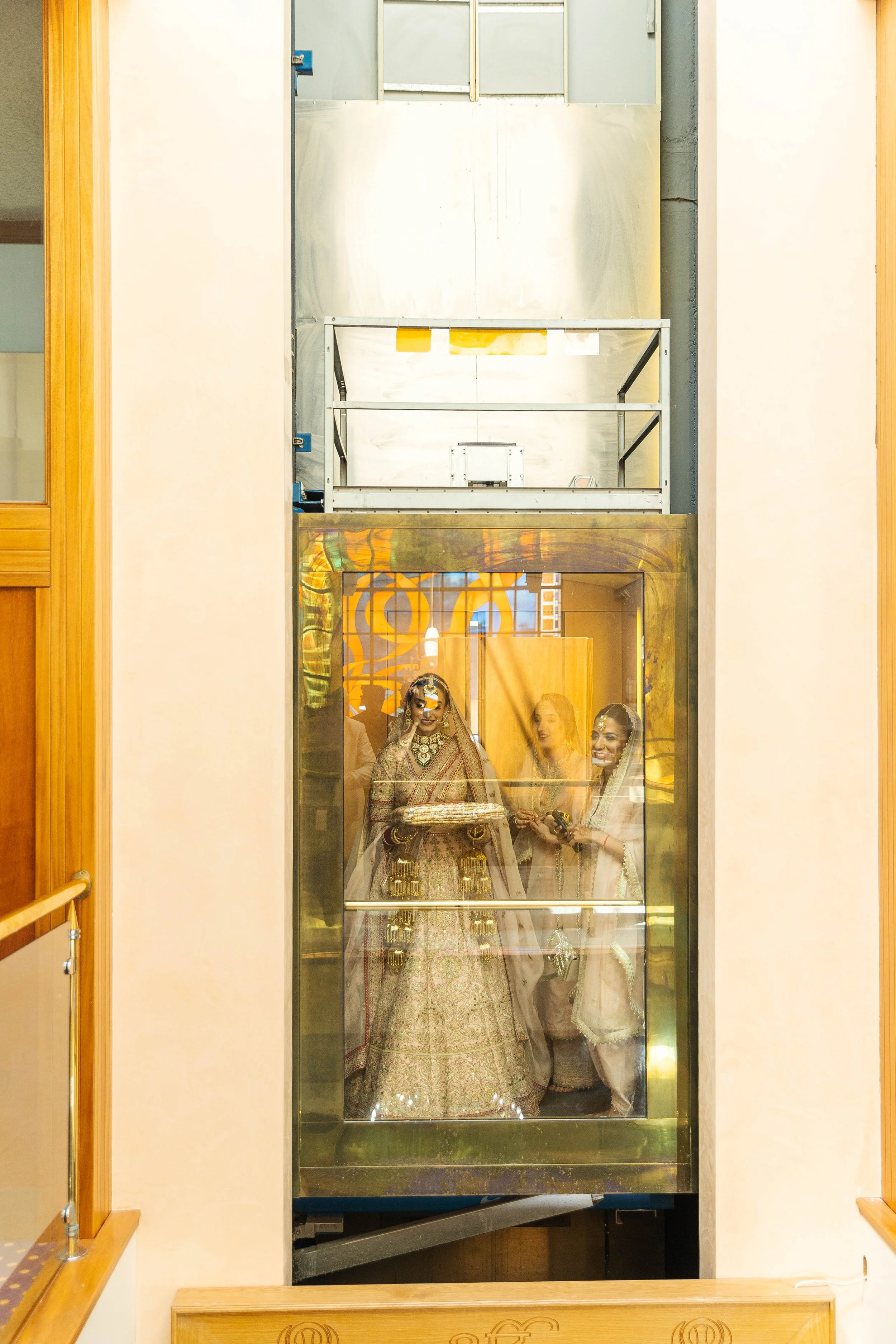 Two women dressed in traditional Indian wedding attire inside an elevator with a glass door, seen from the outside. They are smiling and holding a small item, with a decorated background visible through the glass.