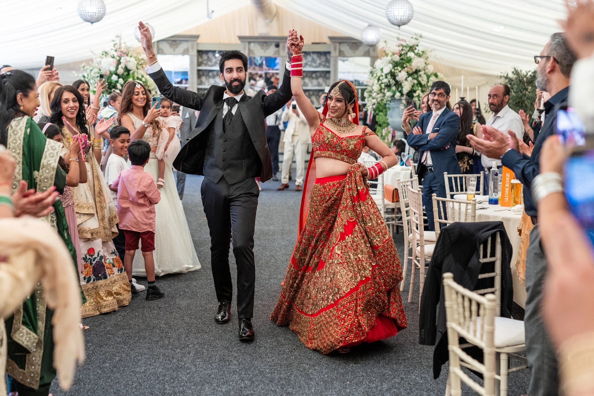 A wedding celebration with a groom and bride dancing inside a decorated venue, surrounded by guests clapping and taking photos.
