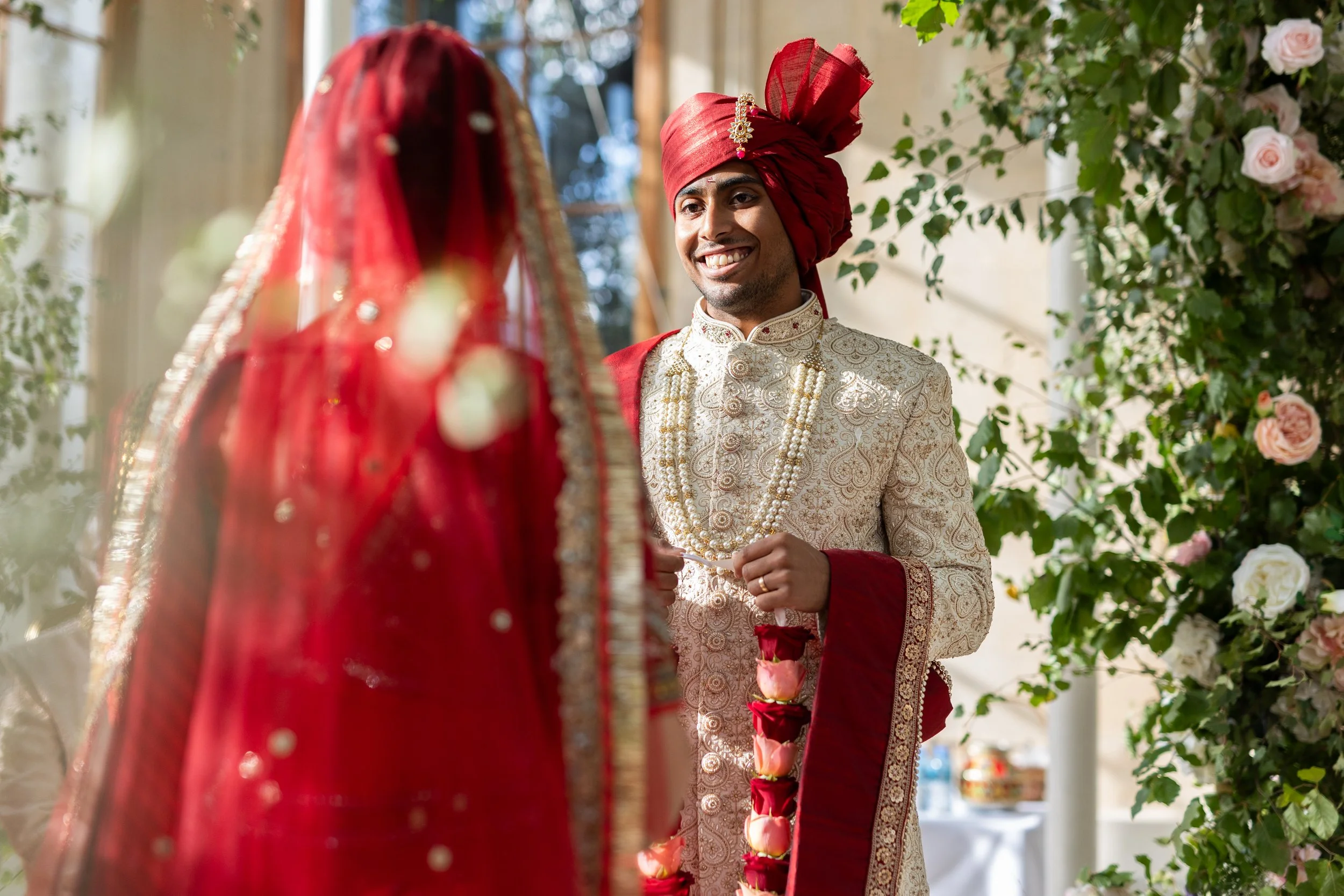 Indian groom wearing traditional attire, including a cream-colored embroidered sherwani and a red turban, smiling at the bride during a wedding ceremony, with floral decorations in the background.