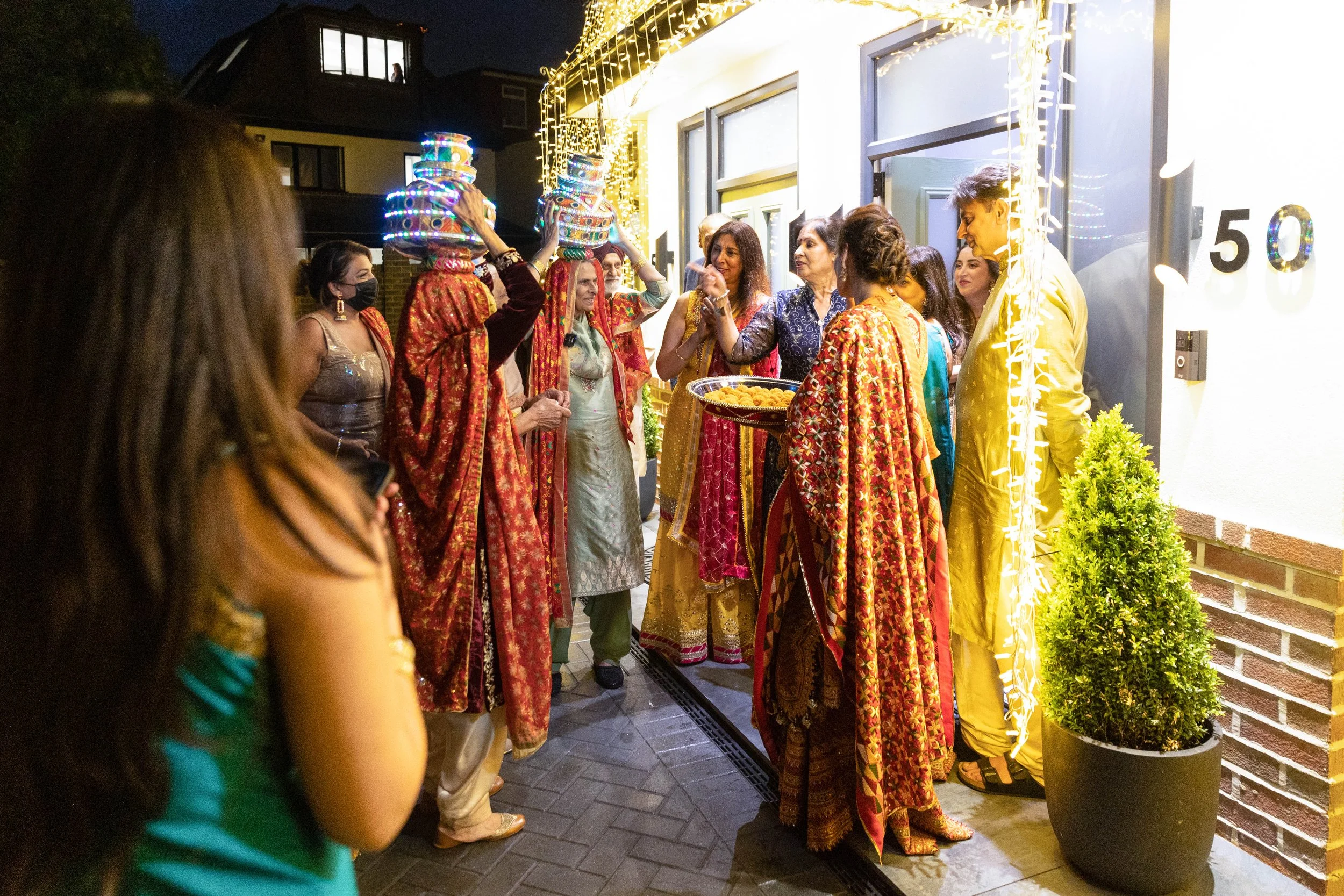 Group of people celebrating during a party outside a house decorated with string lights at night. Some women are wearing traditional Indian sarees, and two individuals are balancing decorated pots on their heads. A woman in a green saree is holding a