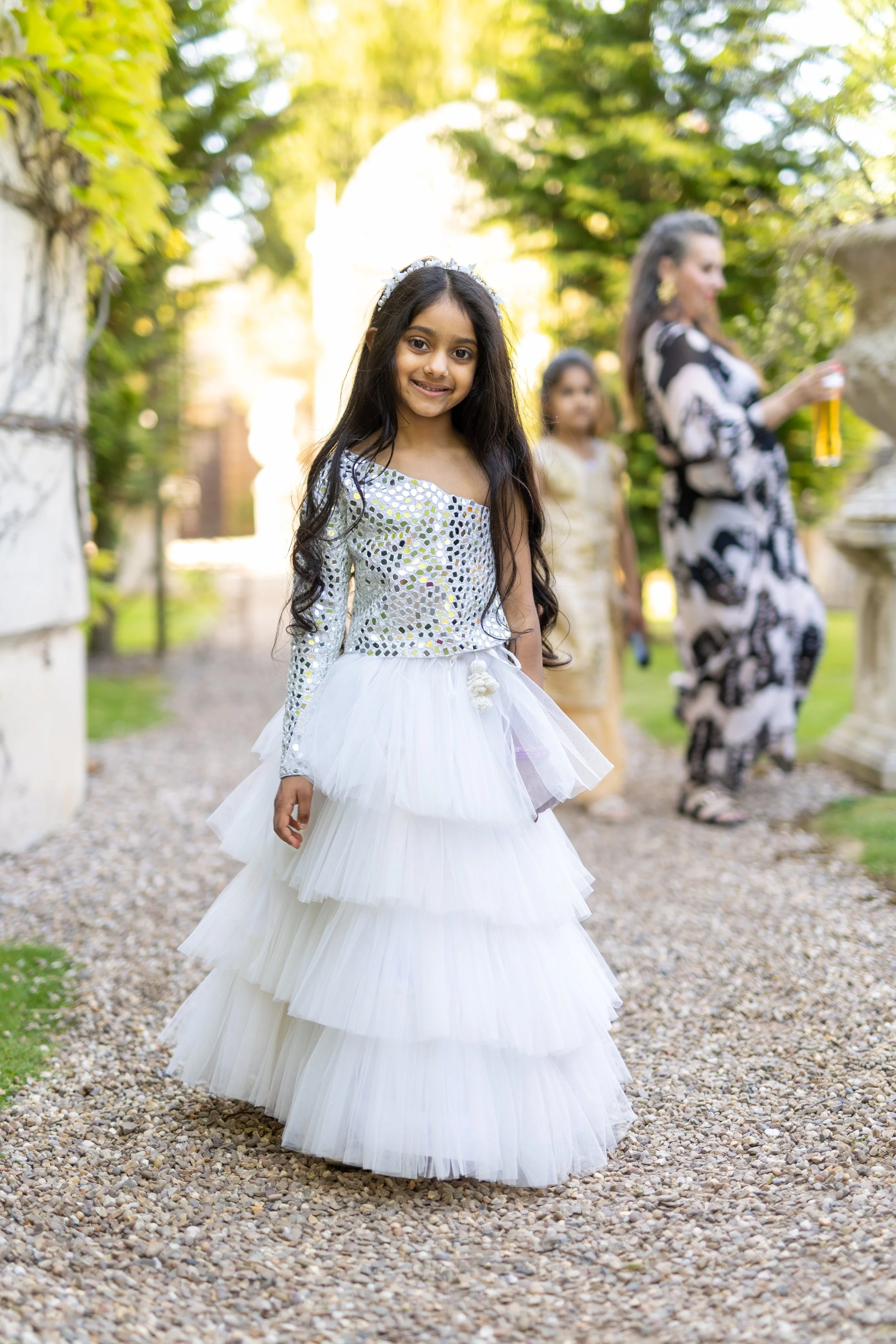 Young girl in a white ball gown with a long tiered tulle skirt, smiling outdoors with greenery, accompanied by two women and another girl in the background.