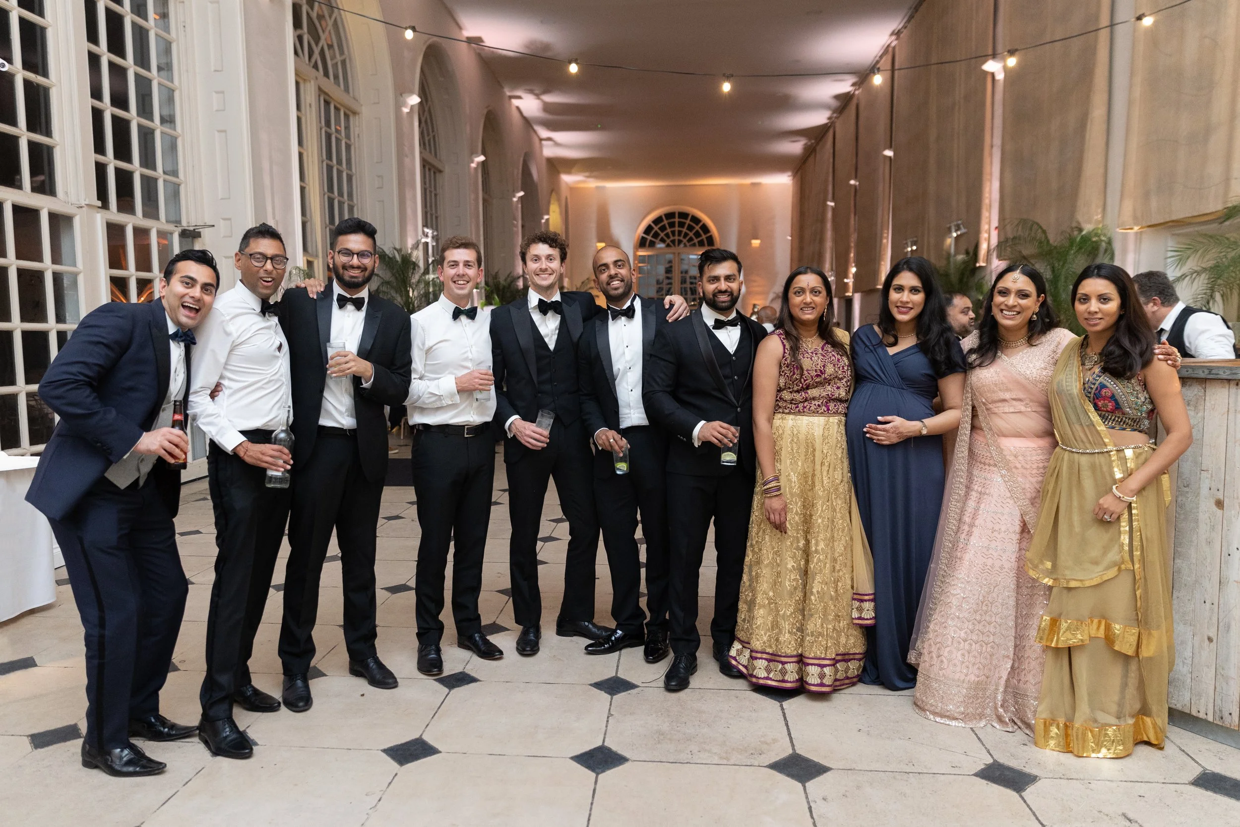 A group of thirteen people, including men in tuxedos and women in colorful dresses, posing together at an indoor event. They are smiling and holding drinks, with a well-lit elegant hall with large windows and string lights in the background.