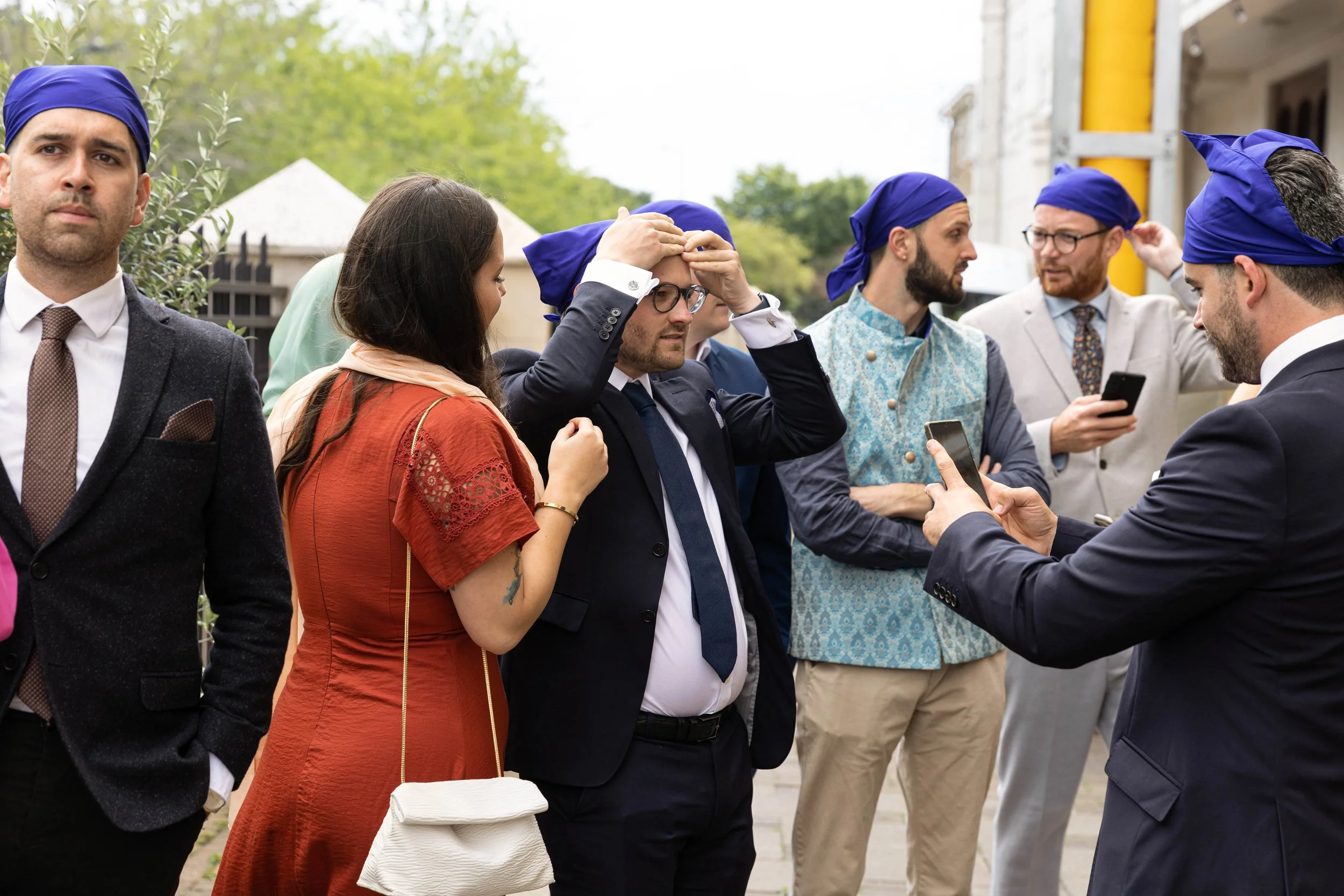 Group of people outside wearing blue head wraps, some using smartphones, with one man adjusting his head wrap.