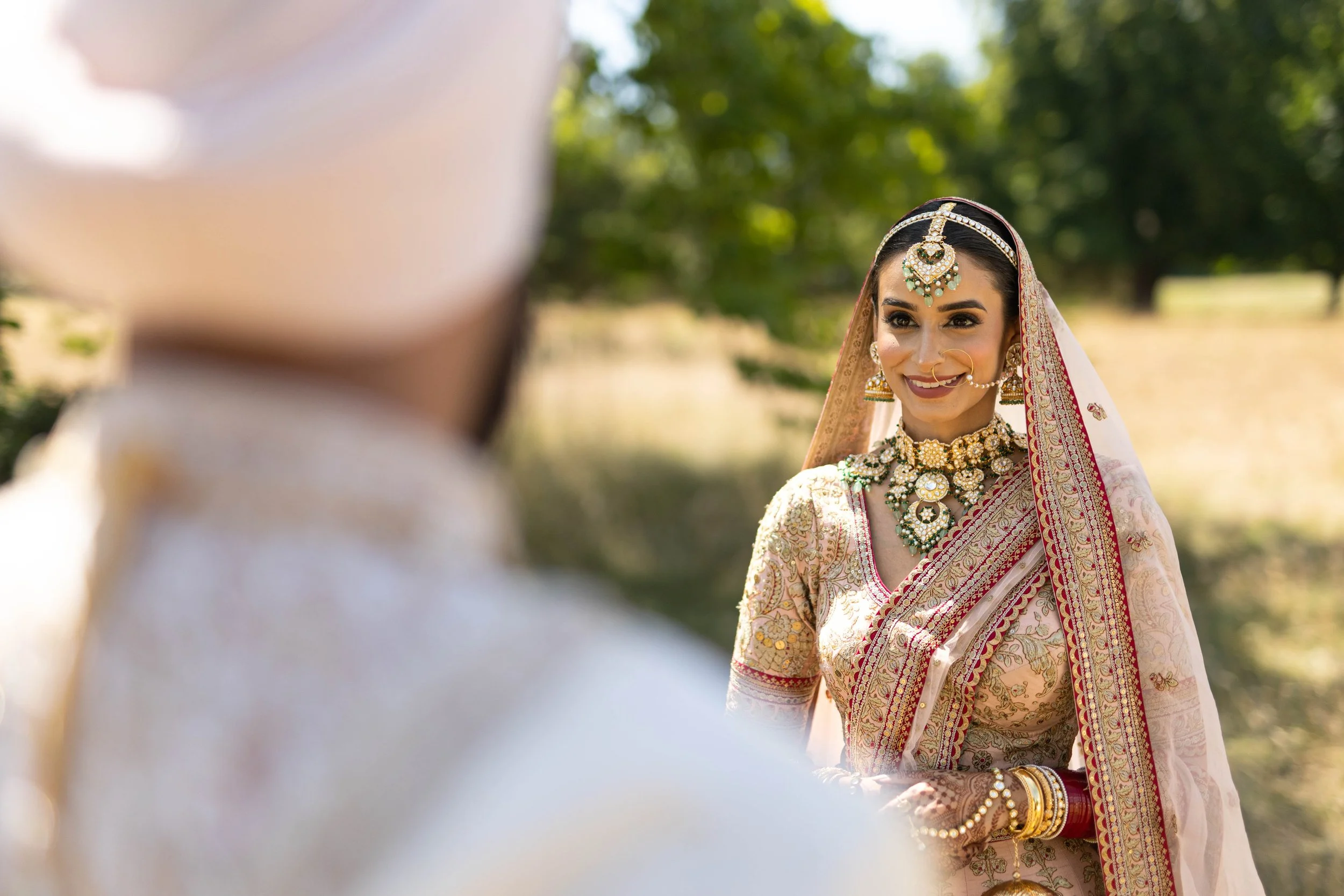 A woman dressed in traditional Indian attire with elaborate jewelry and a saree, smiling at a man whose back is visible in the foreground, outdoors with green trees in the background.
