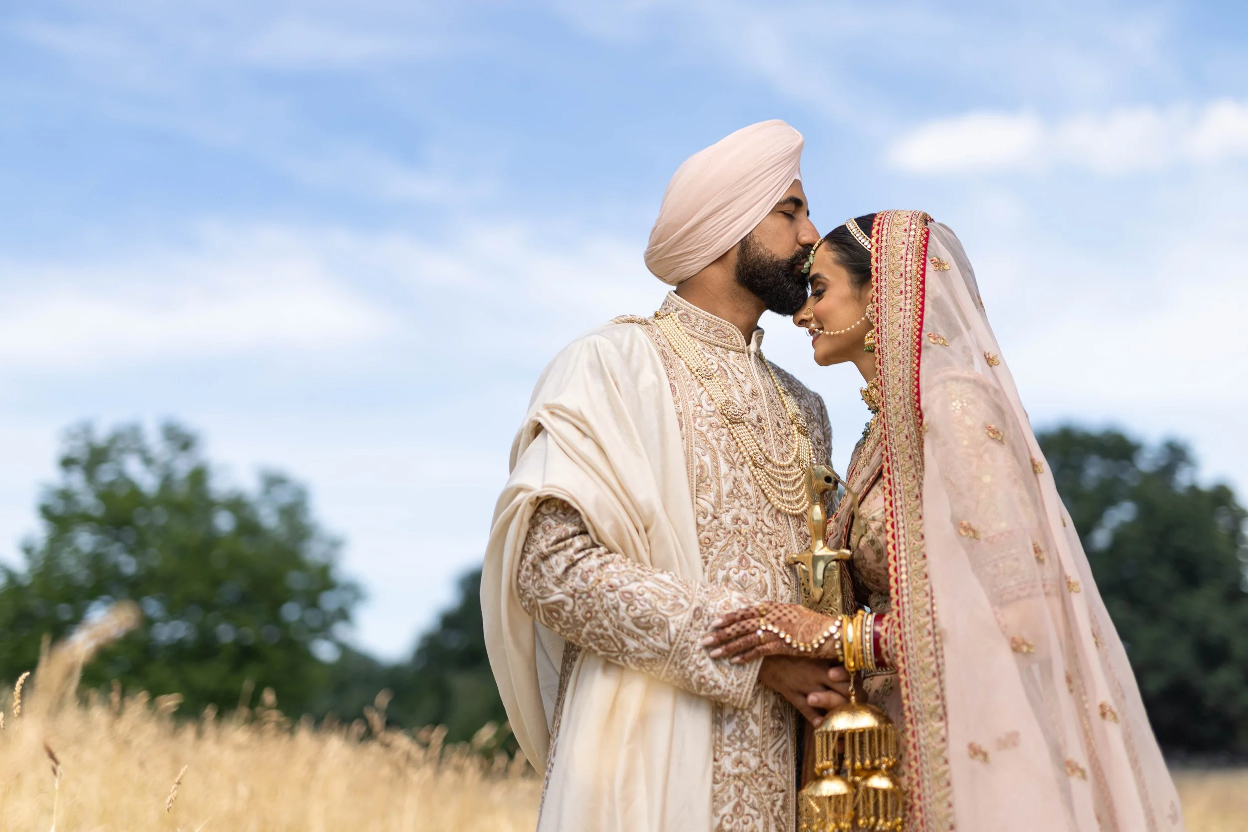 An Indian couple in traditional wedding attire standing outdoors in a field, sharing an intimate moment with foreheads touching, with trees and a cloudy sky in the background.