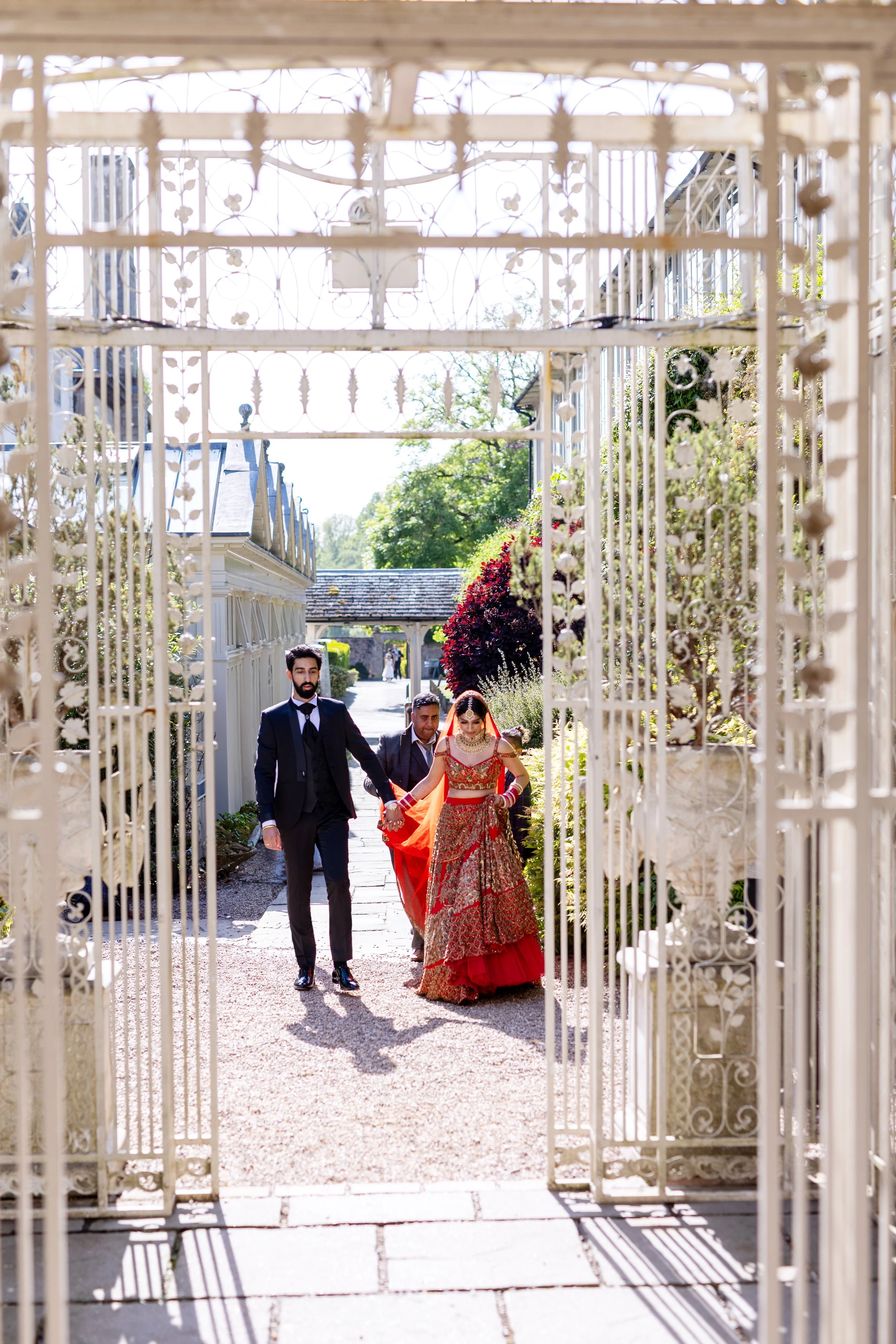 A bride dressed in a red and gold traditional Indian wedding attire, holding hands with a groom in a black suit, walking through a white ornate gate outdoors on a sunny day.