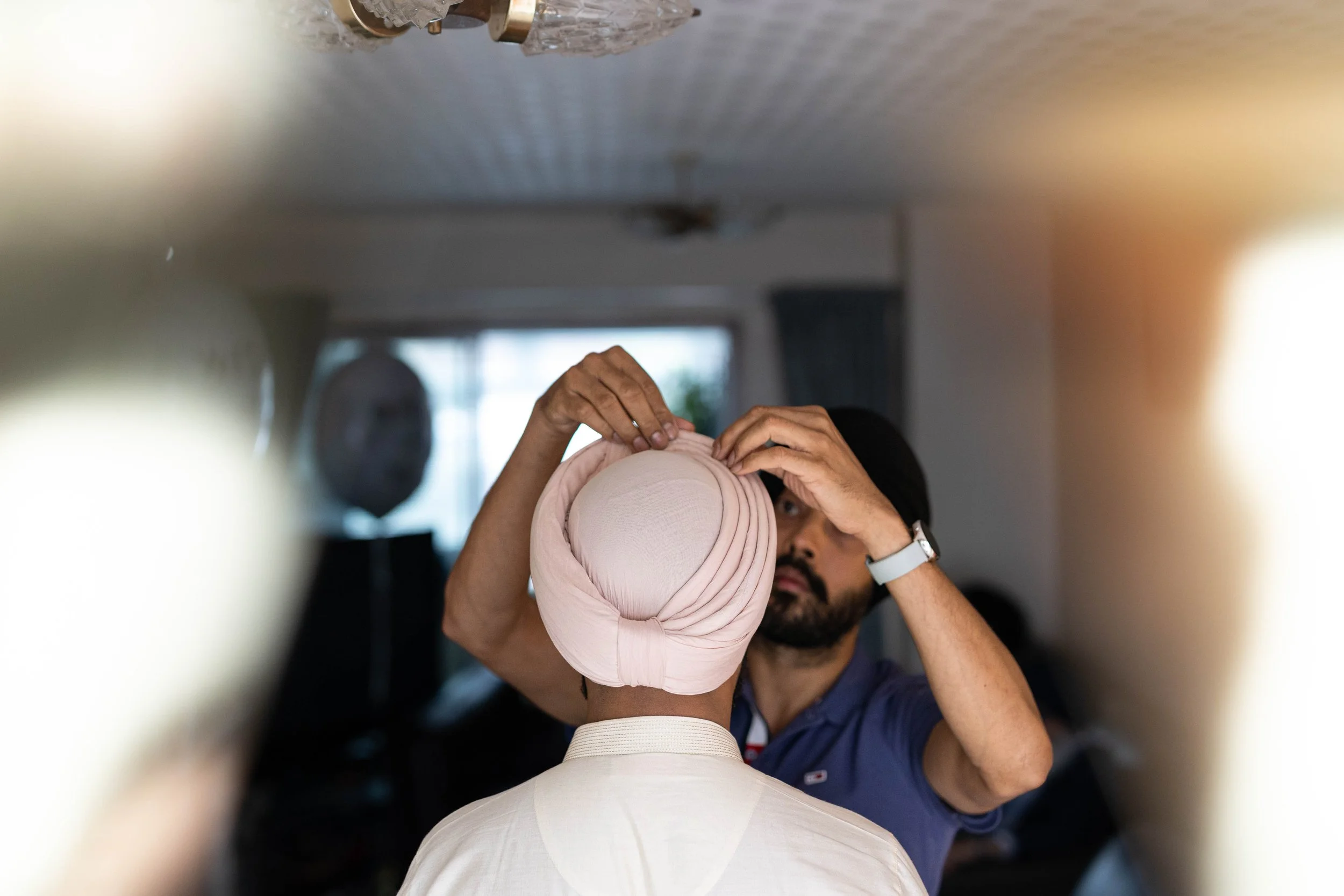 A person adjusting a light pink headscarf on another person's head in a room with a window and a speaker in the background.