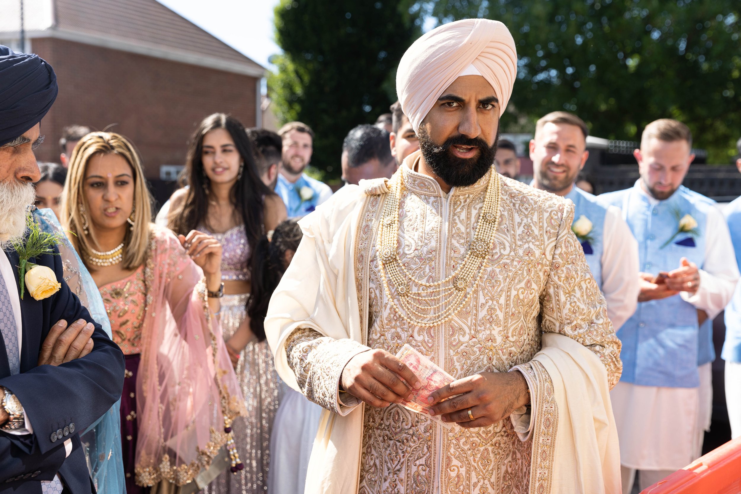 A groom of Indian ethnicity at a traditional wedding ceremony, dressed in an ornate cream-colored sherwani and turban, holding money, surrounded by friends and family in colorful traditional attire.
