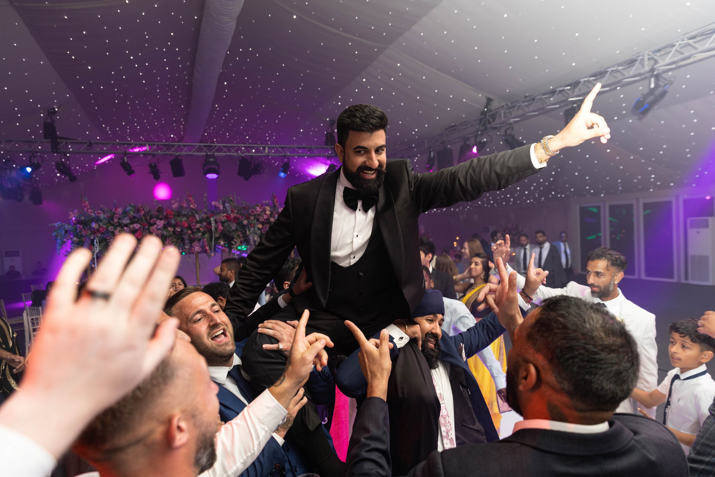 A man in a black tuxedo with a bowtie is being lifted by others at a celebration or wedding reception. The man is smiling and pointing upwards. The background features purple and pink lighting, floral arrangements, and other guests.