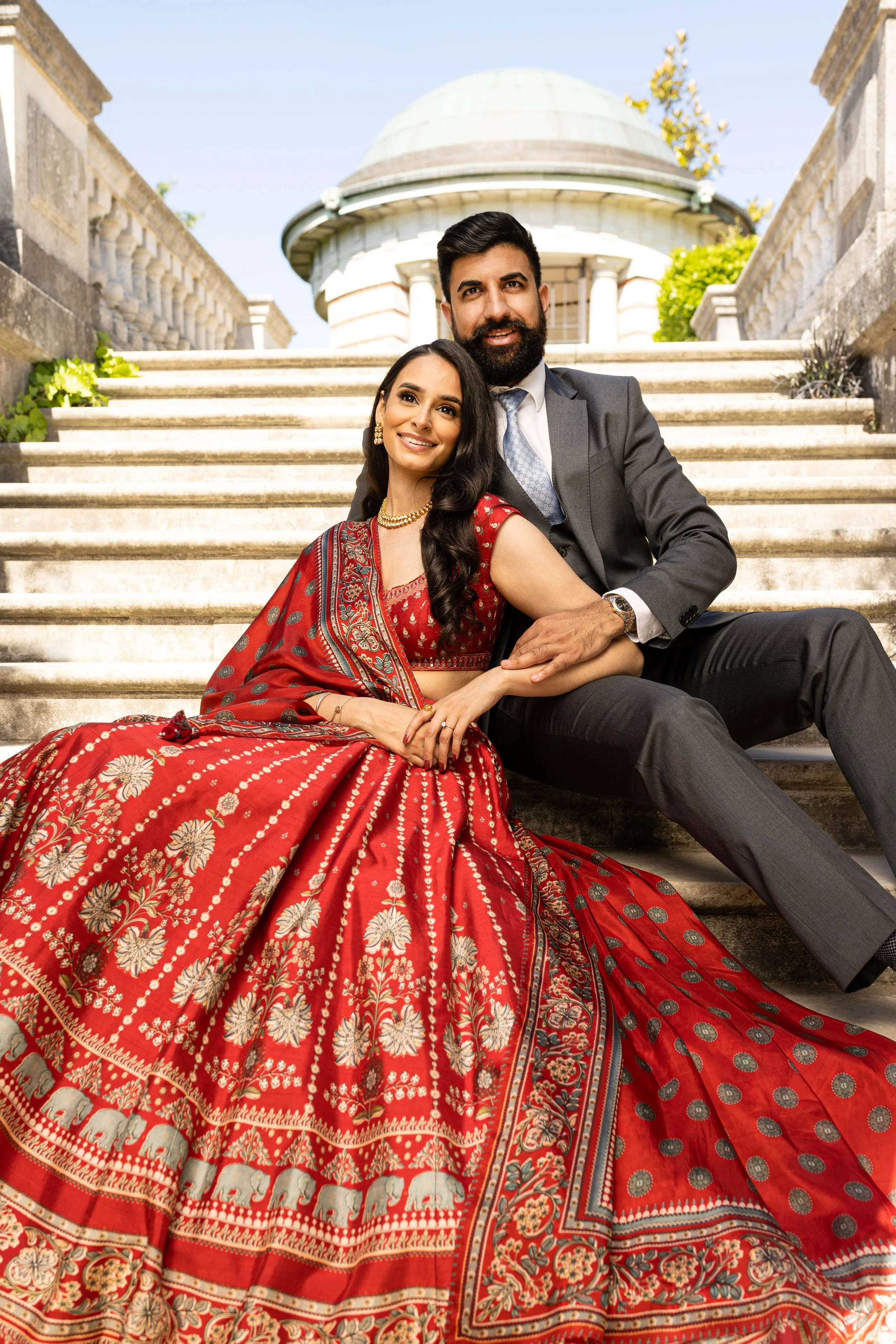 A couple sitting on steps outside a historic building, featuring a woman in a red traditional Indian dress and a man in a suit, under clear blue sky.