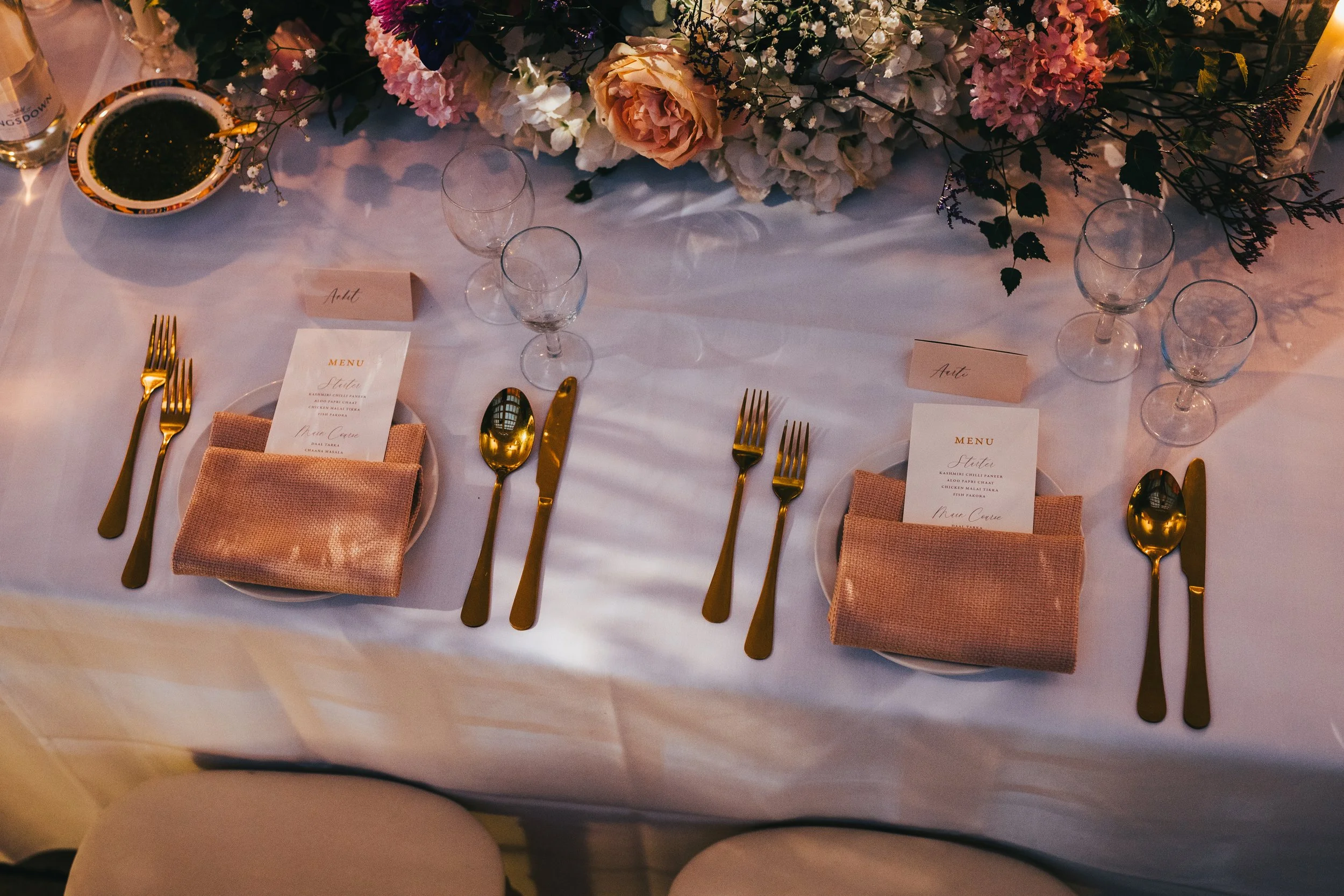 Elegant dining table setup with gold flatware, pink napkins on white plates, water glasses, and floral centerpiece with roses and hydrangeas.