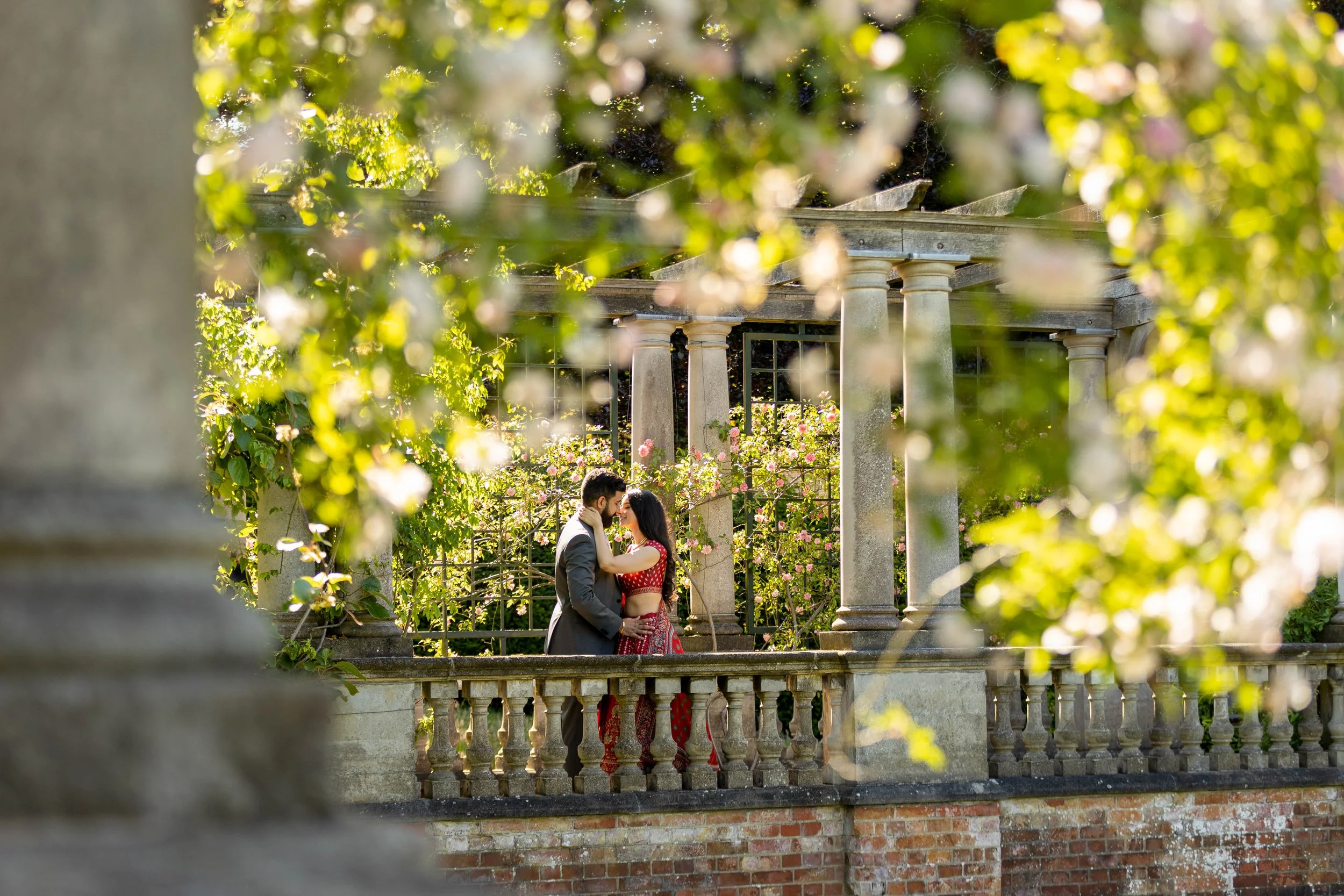 A couple dressed in formal attire sharing a romantic moment on a stone balcony surrounded by lush greenery and blooming flowers, with classical columns in the background.