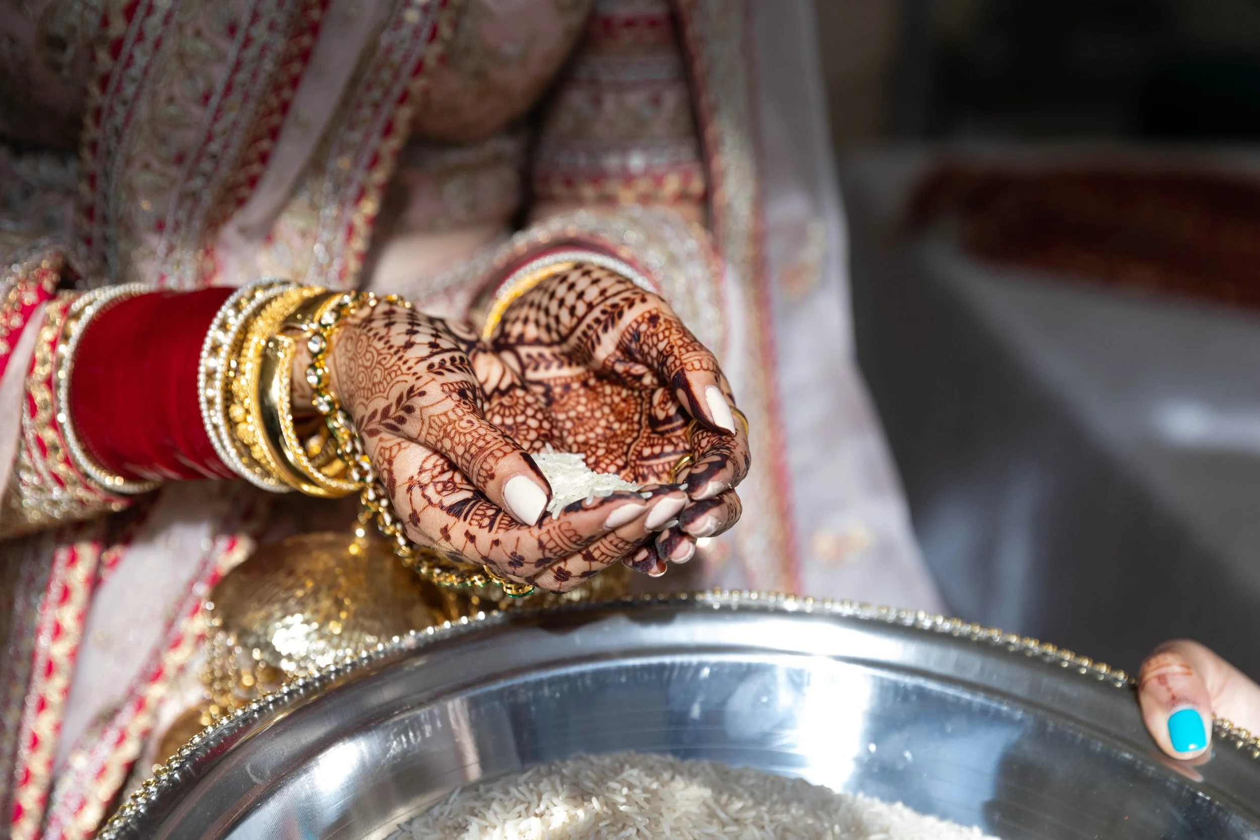 A woman with intricate henna designs on her hands, wearing traditional jewelry and red and gold attire, holding a small piece of white material over a large bowl of rice.