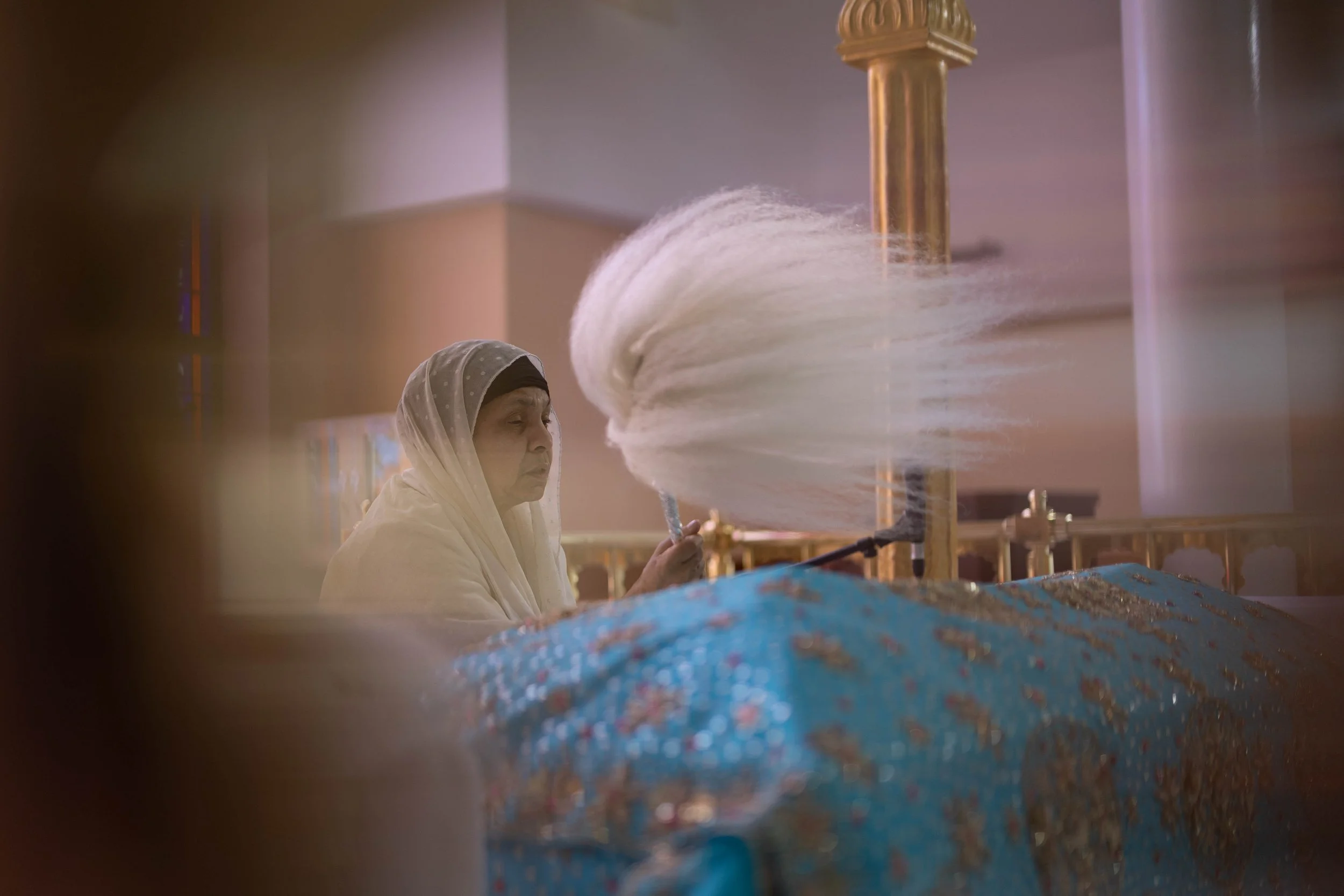 An elderly woman dressed in traditional attire is sitting inside a mosque, observing a large chandelier being cleaned or maintained. The chandelier has long, white, flowing details and is suspended from the ceiling. The scene suggests a moment of qui