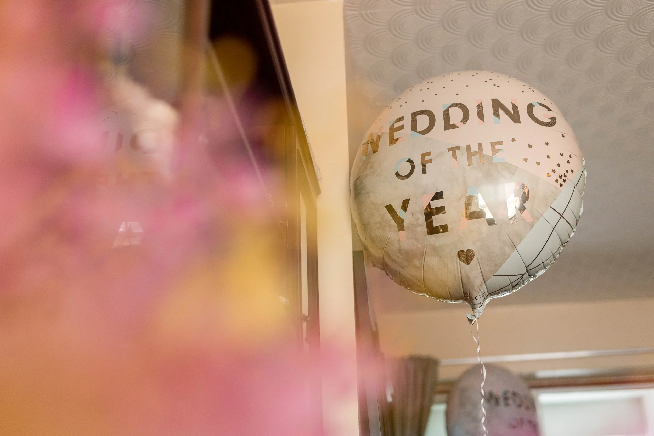 A round balloon with the text "WEDDING OF THE YEAR" printed on it, decorated with small black hearts and colorful triangles, floating in a room with pink and yellow flowers in the foreground.